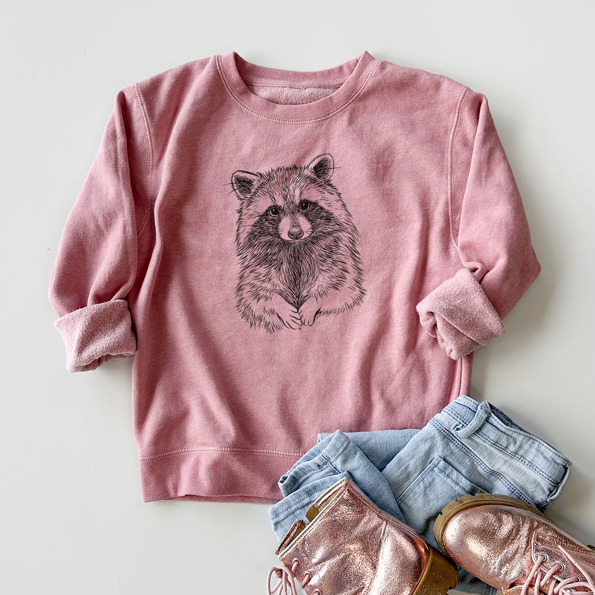 Youth crewneck sweatshirt printed with a detailed black ink portrait of a raccoon with its front paws resting in front of it, styled flat with jeans and boots.