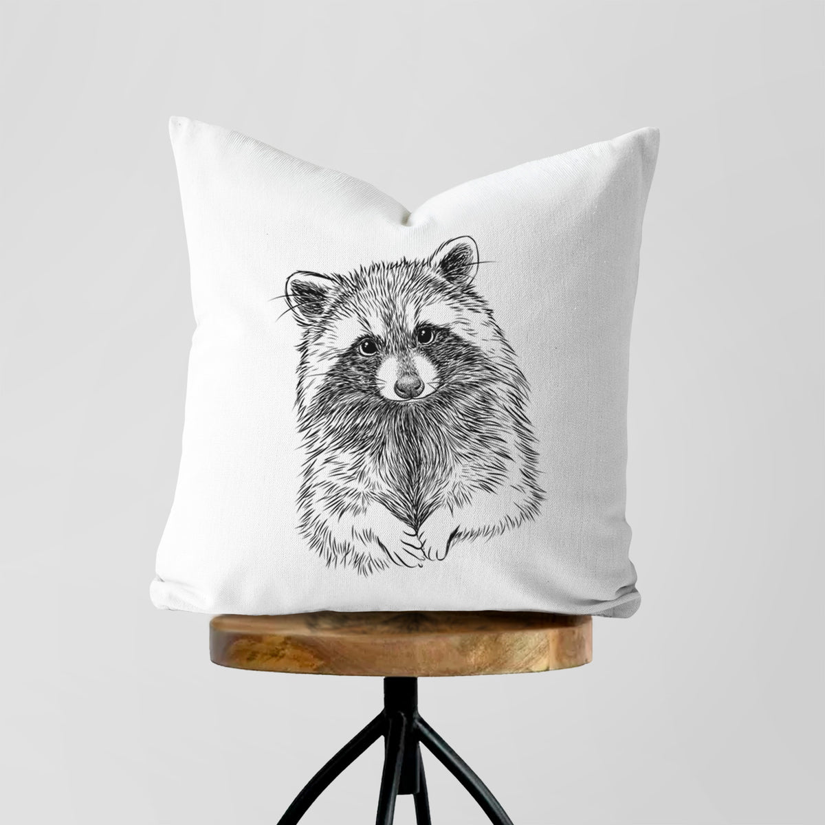 White pillow cover printed with a detailed black ink portrait of a raccoon with its front paws resting in front of it, on a wooden stool.