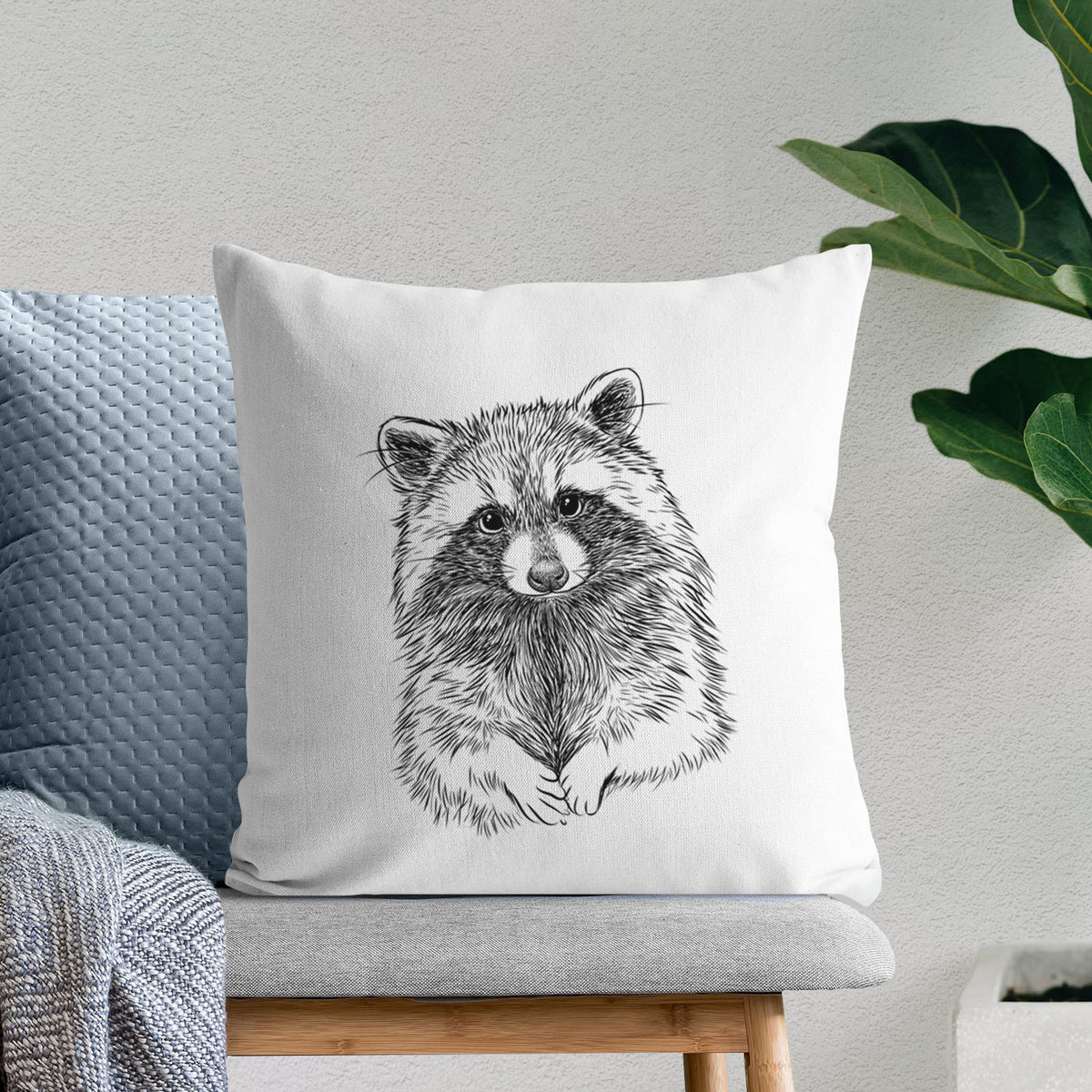 White pillow cover printed with a detailed black ink portrait of a raccoon with its front paws resting in front of it, on a bench beside plants.