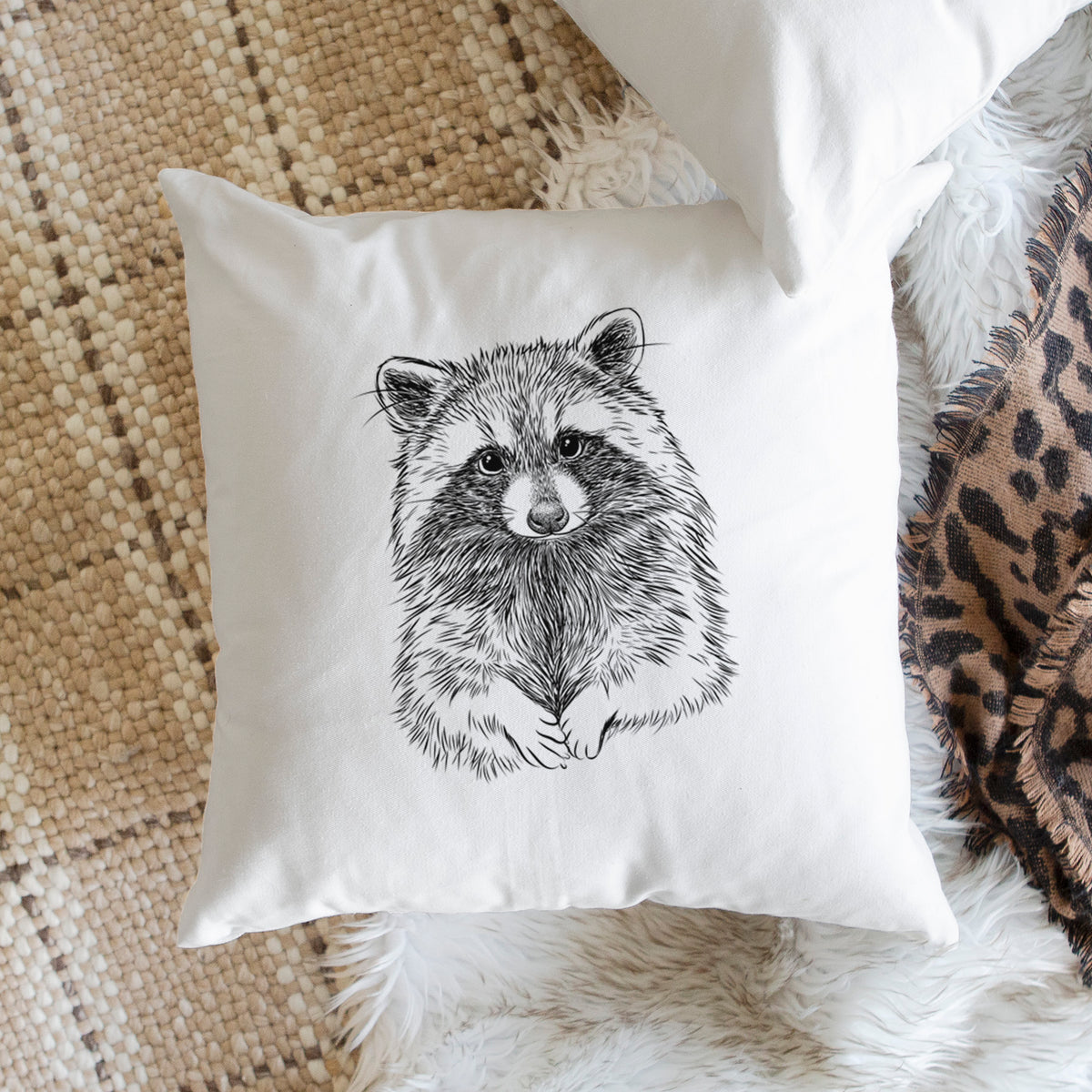 White pillow cover printed with a detailed black ink portrait of a raccoon with its front paws resting in front of it, on a woven chair.