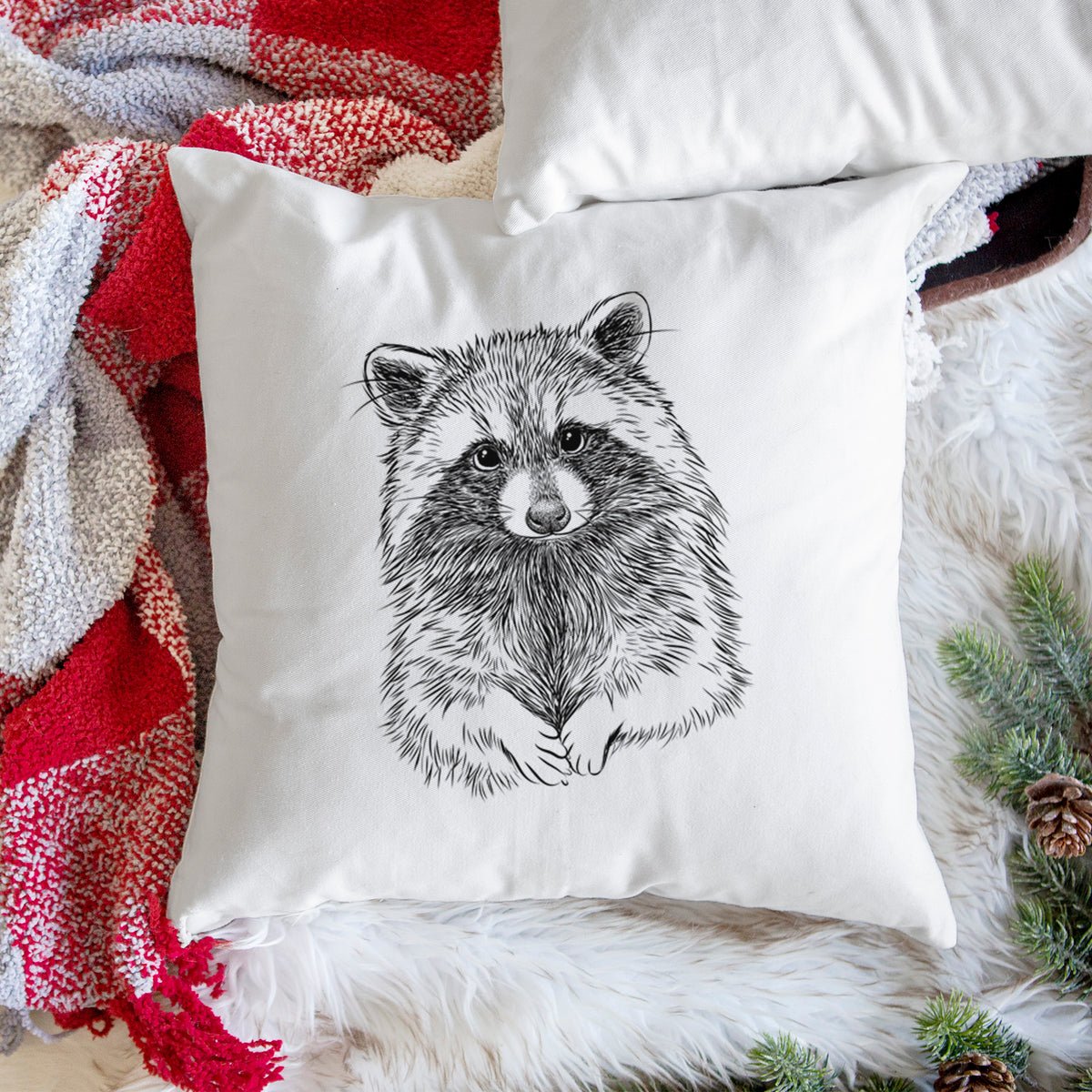 White pillow cover printed with a detailed black ink portrait of a raccoon with its front paws resting in front of it, on a woven chair.