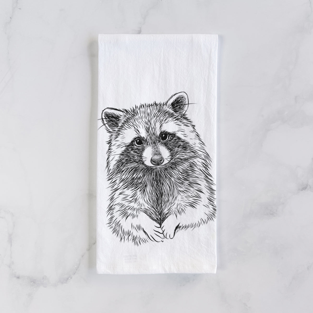White cotton tea towel laid flat on marble, printed with a detailed black ink portrait of a raccoon with its front paws resting in front of it.