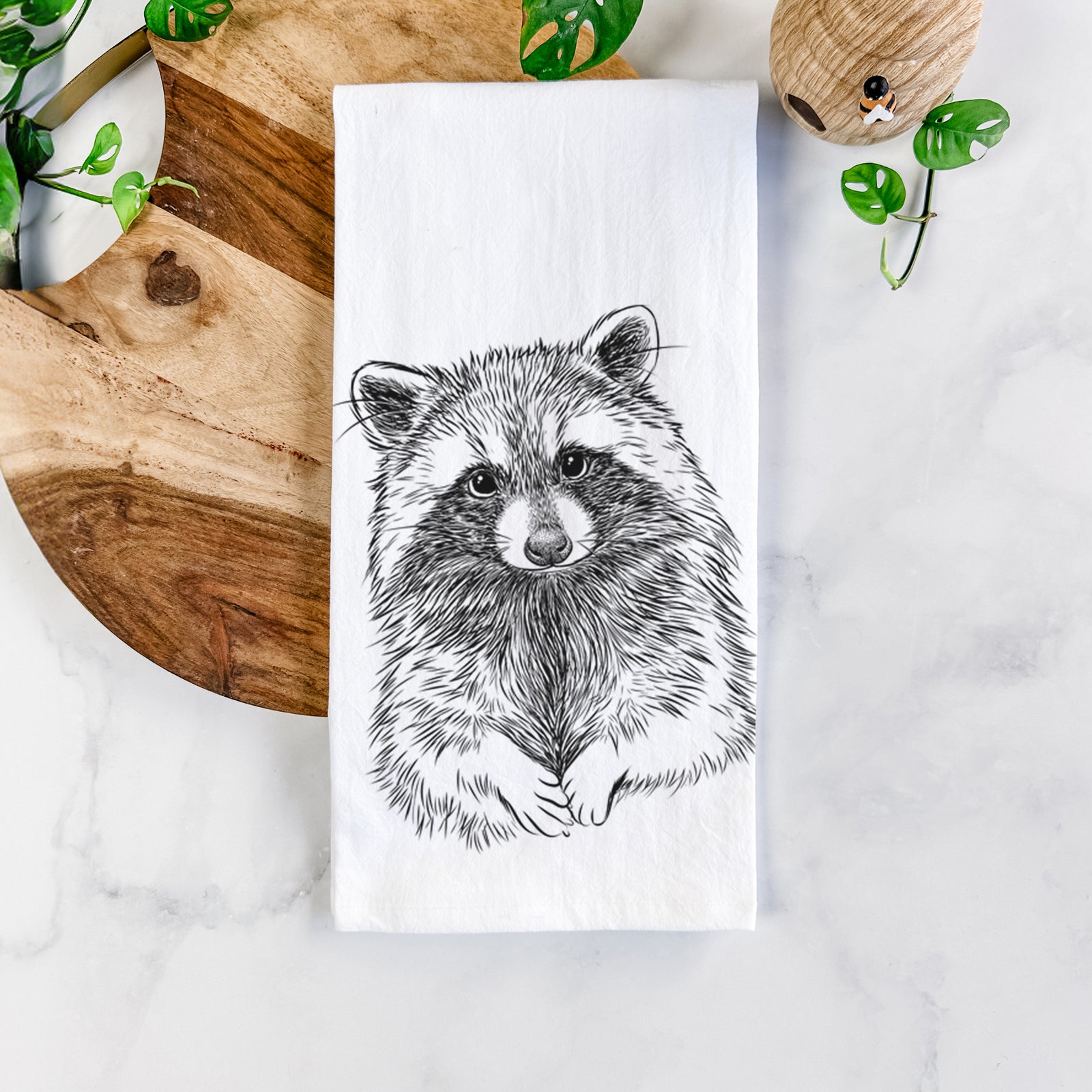 White cotton tea towel printed with a detailed black ink portrait of a raccoon with its front paws resting in front of it, on a marble counter beside a round wooden cutting board and greenery.