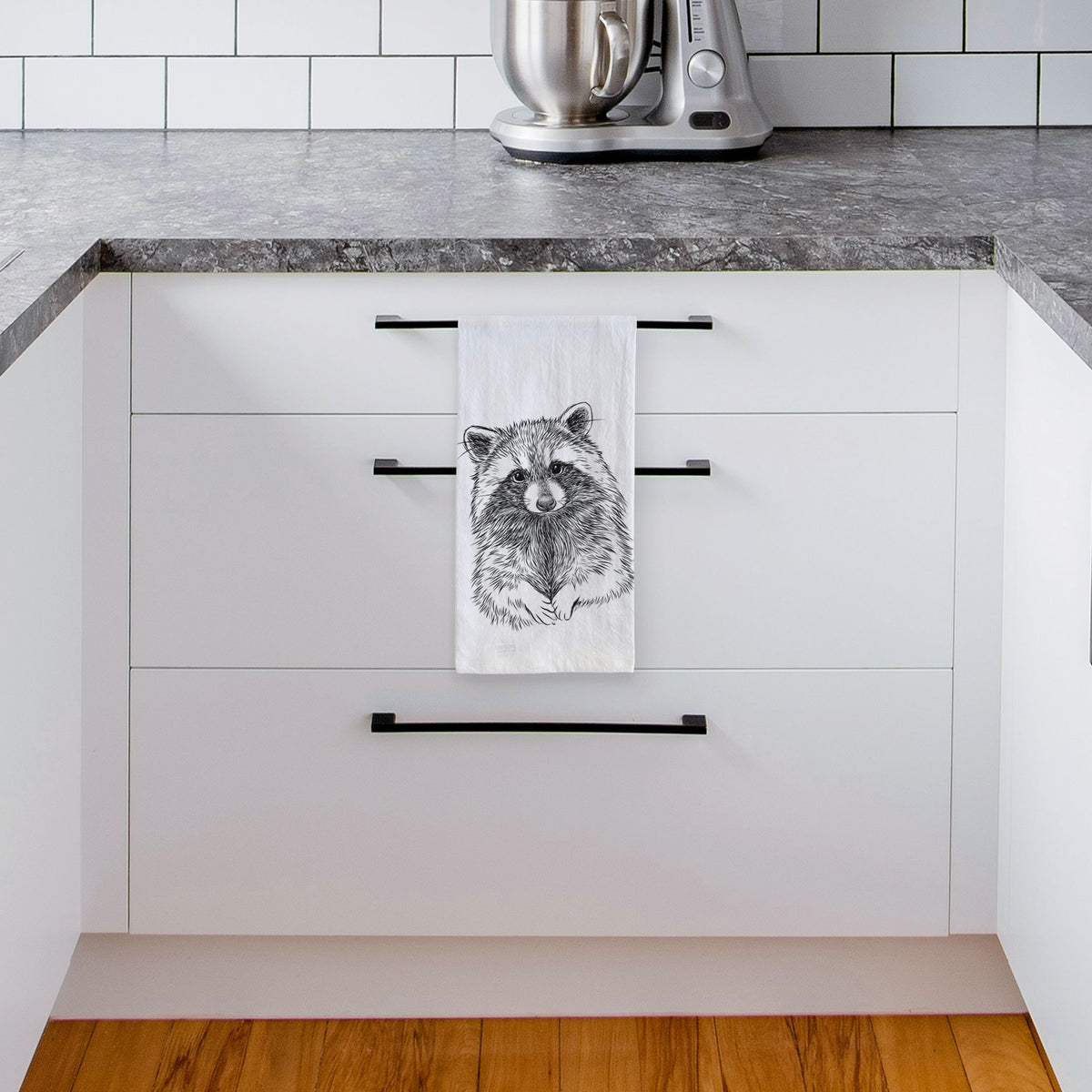 White tea towel printed with a detailed black ink portrait of a raccoon with its front paws resting in front of it, hanging from a drawer handle in a bright white kitchen.
