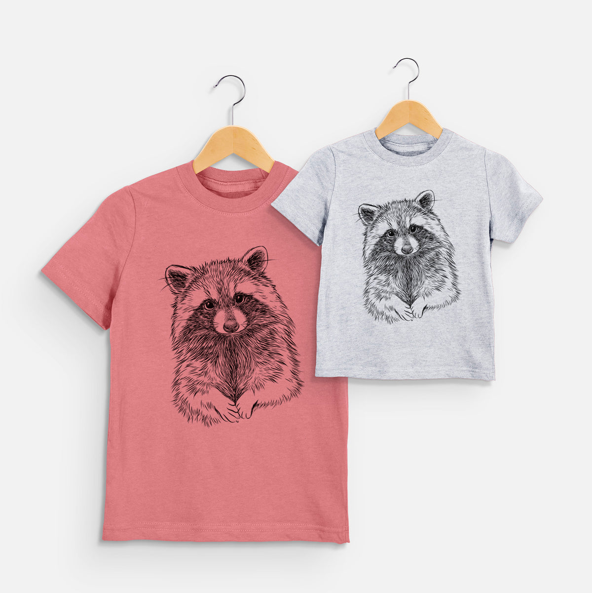 Two youth t-shirts in different colors on hangers, printed with a detailed black ink portrait of a raccoon with its front paws resting in front of it.