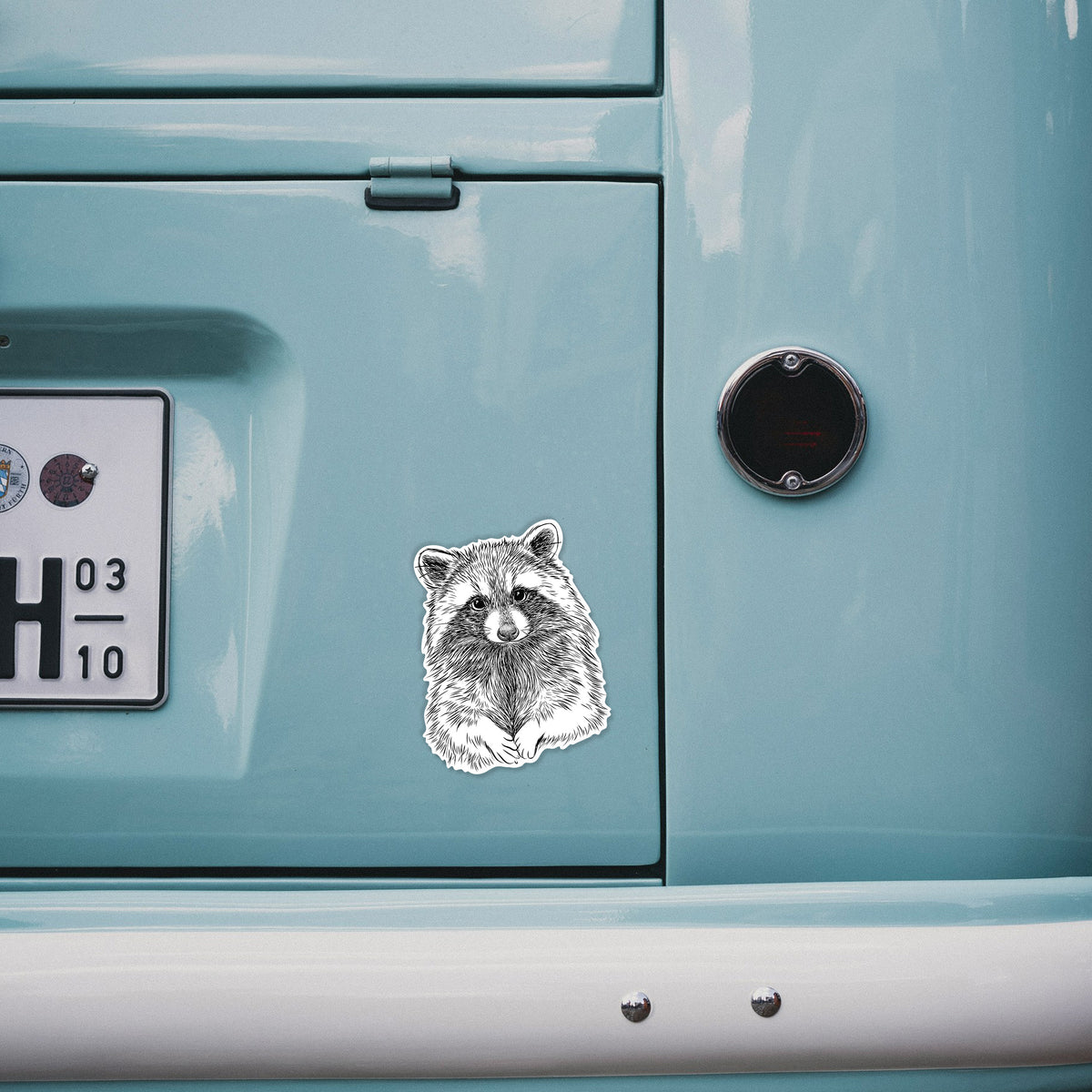 4 inch vinyl decal of a detailed black ink portrait of a raccoon with its front paws resting in front of it on the back of a van.