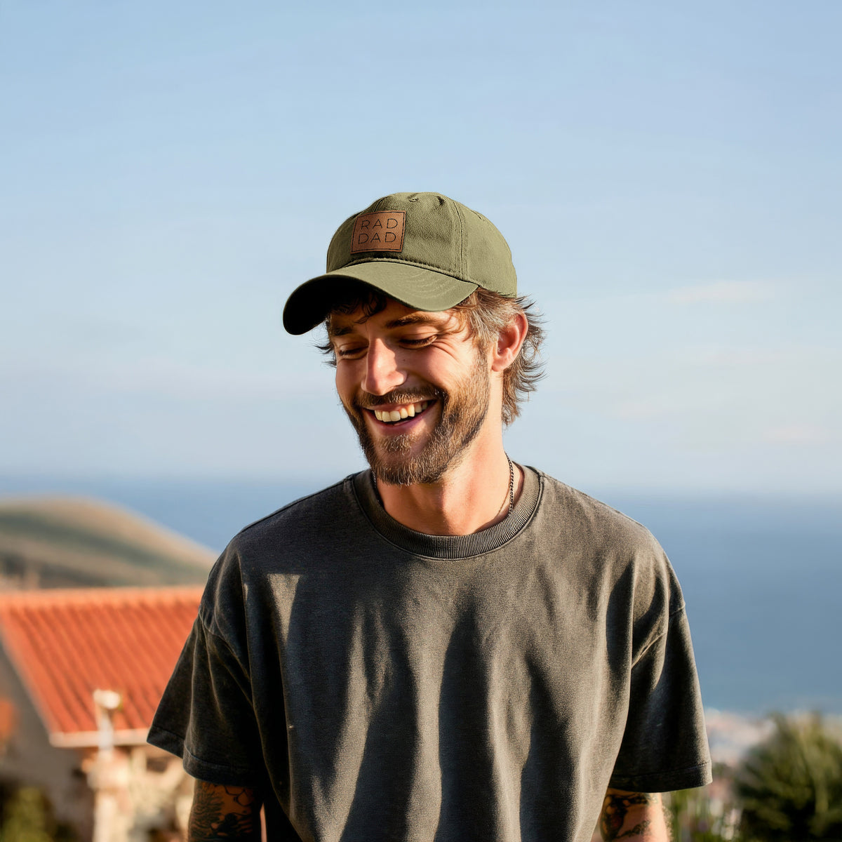 Smiling man outdoors near the ocean wearing an olive green baseball hat and a dark t-shirt.