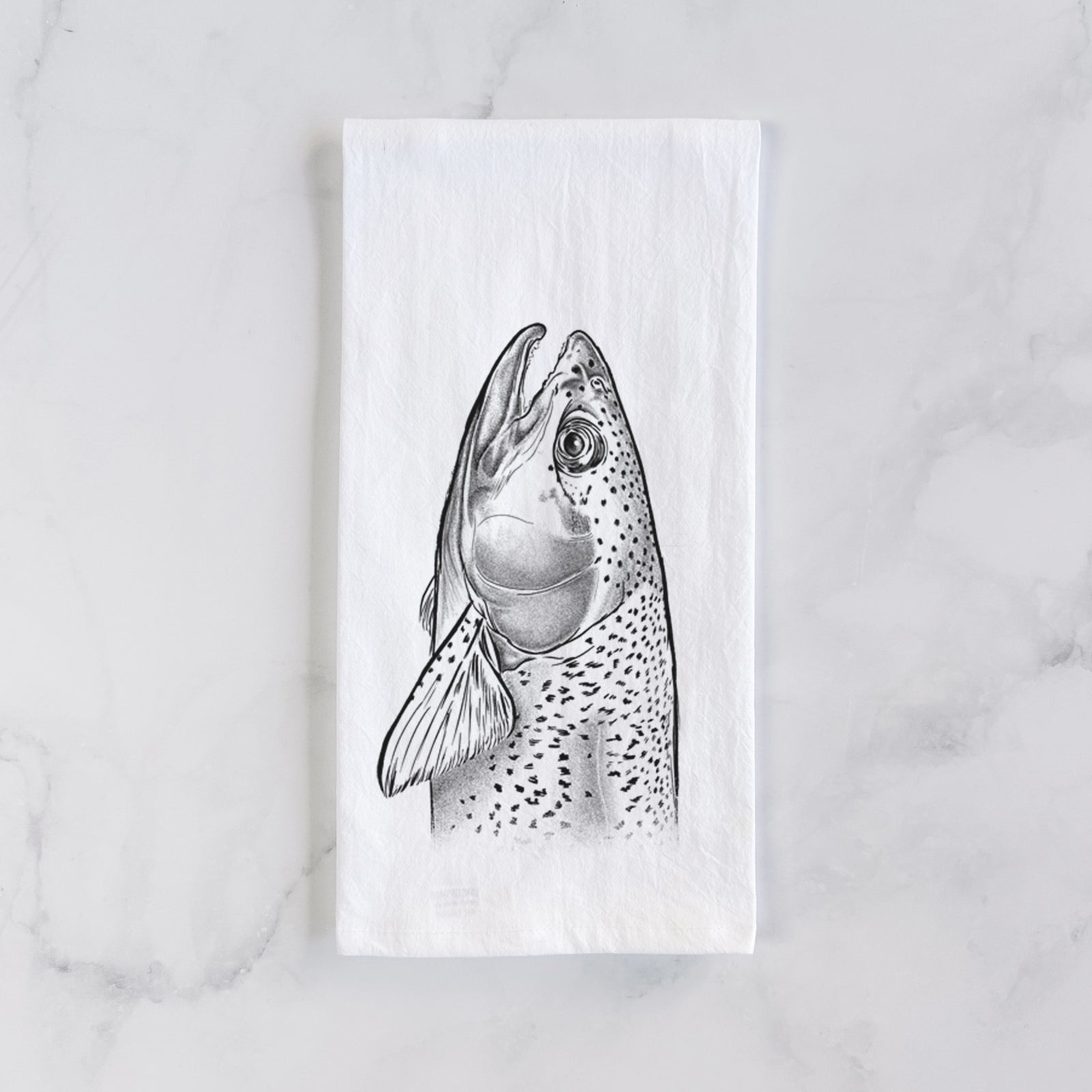 White cotton tea towel printed with a detailed black ink illustration of a rainbow trout's head pointing upward, showing its spotted scales, on a marble counter beside a round wooden cutting board and greenery.