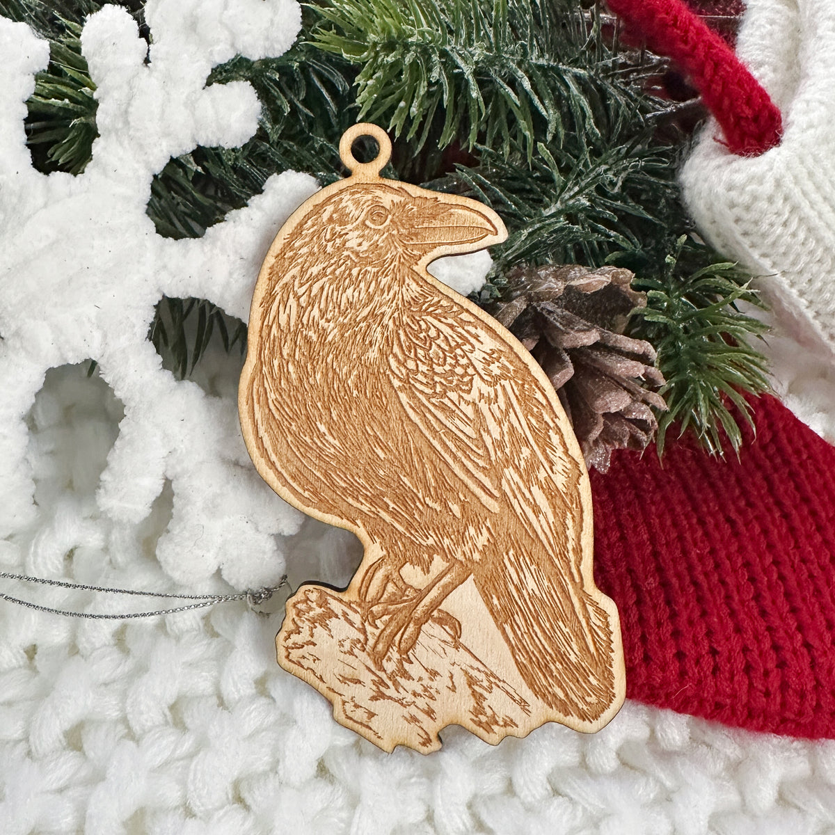 Wooden ornament featuring a detailed engraving of a raven perched, on a knit blanket with pine cones.
