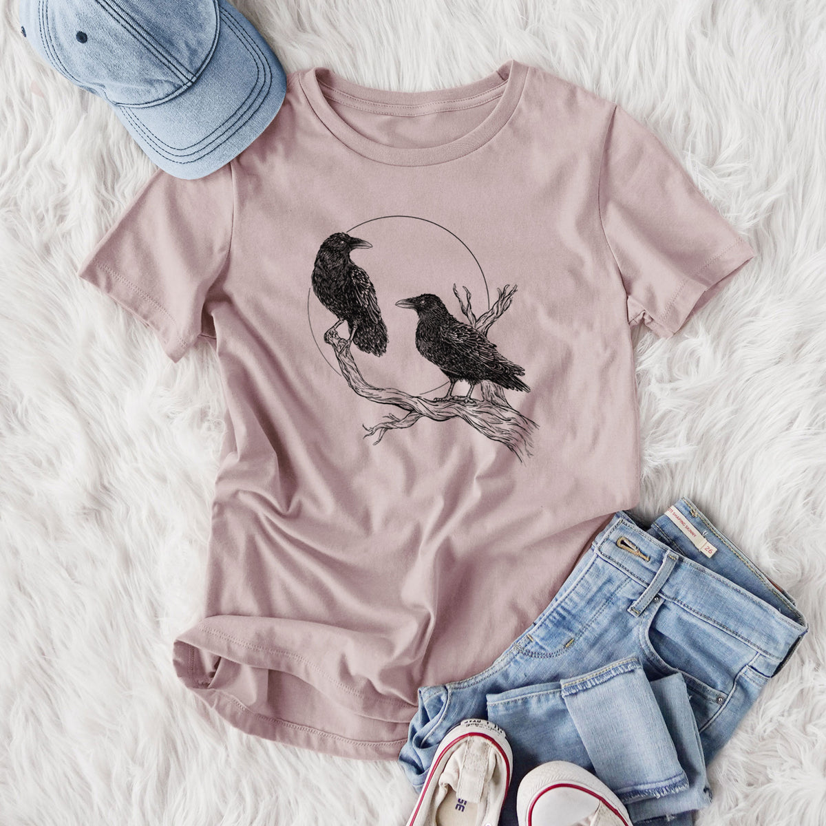 Women's relaxed fit cotton t-shirt printed with a black ink illustration of two ravens perched on a branch in front of a full moon, styled flat on a white faux fur rug with jeans.