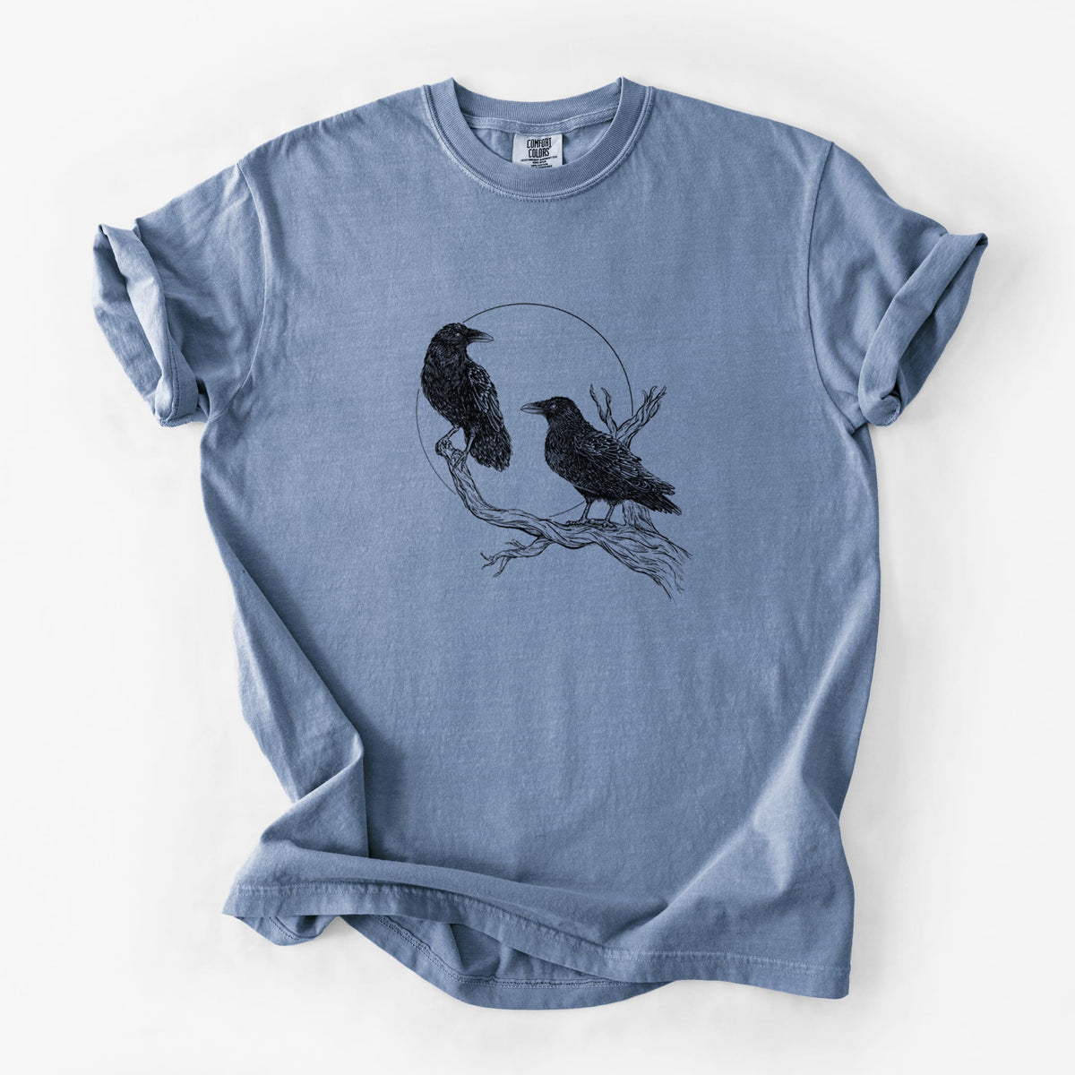 Blue jean Comfort Colors t-shirt laid flat, printed with a black ink illustration of two ravens perched on a branch in front of a full moon.