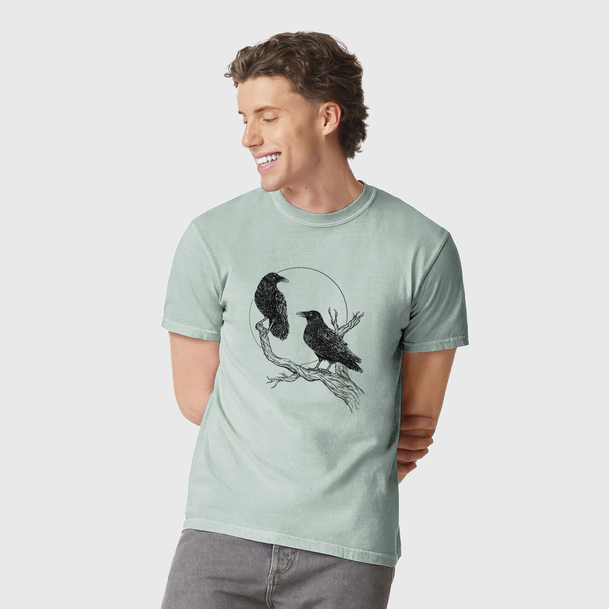 Man wearing a Comfort Colors t-shirt printed with a black ink illustration of two ravens perched on a branch in front of a full moon.