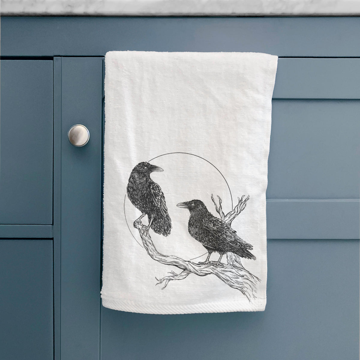 White hand towel printed with a black ink illustration of two ravens perched on a branch in front of a full moon, hanging on a cabinet door.