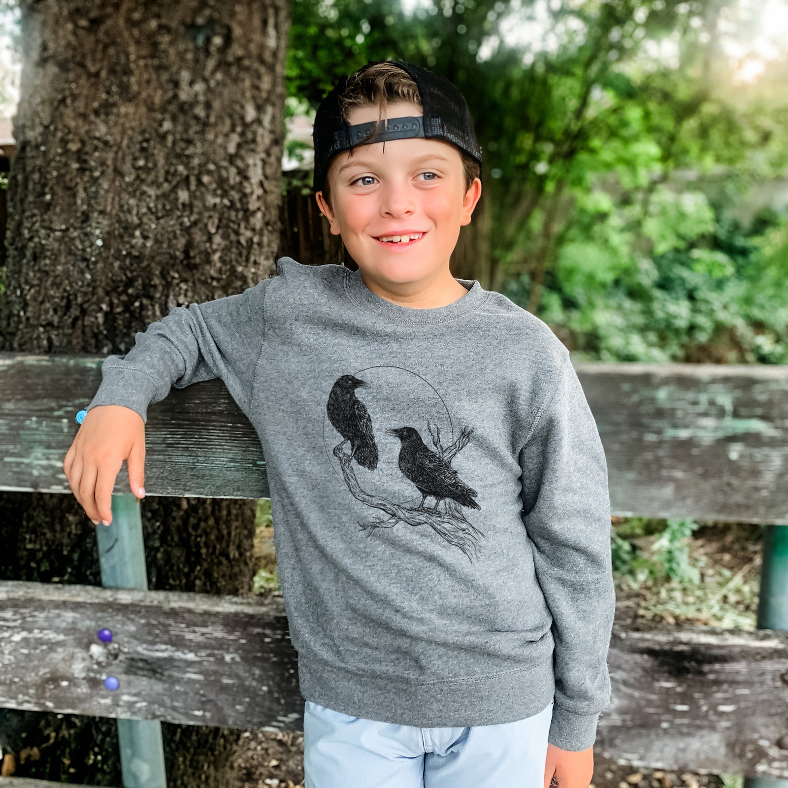Boy wearing a youth crewneck sweatshirt printed with a black ink illustration of two ravens perched on a branch in front of a full moon, outdoors.