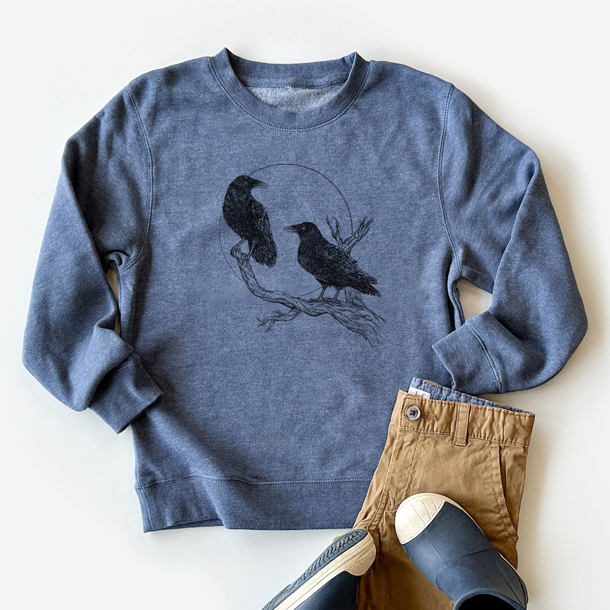 Youth crewneck sweatshirt printed with a black ink illustration of two ravens perched on a branch in front of a full moon, styled flat with khaki pants and shoes.