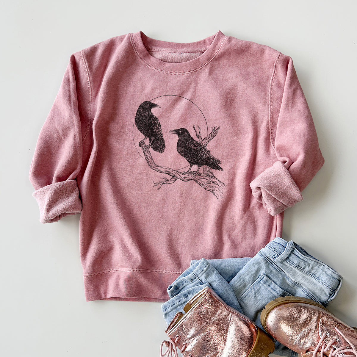 Youth crewneck sweatshirt printed with a black ink illustration of two ravens perched on a branch in front of a full moon, styled flat with jeans and boots.