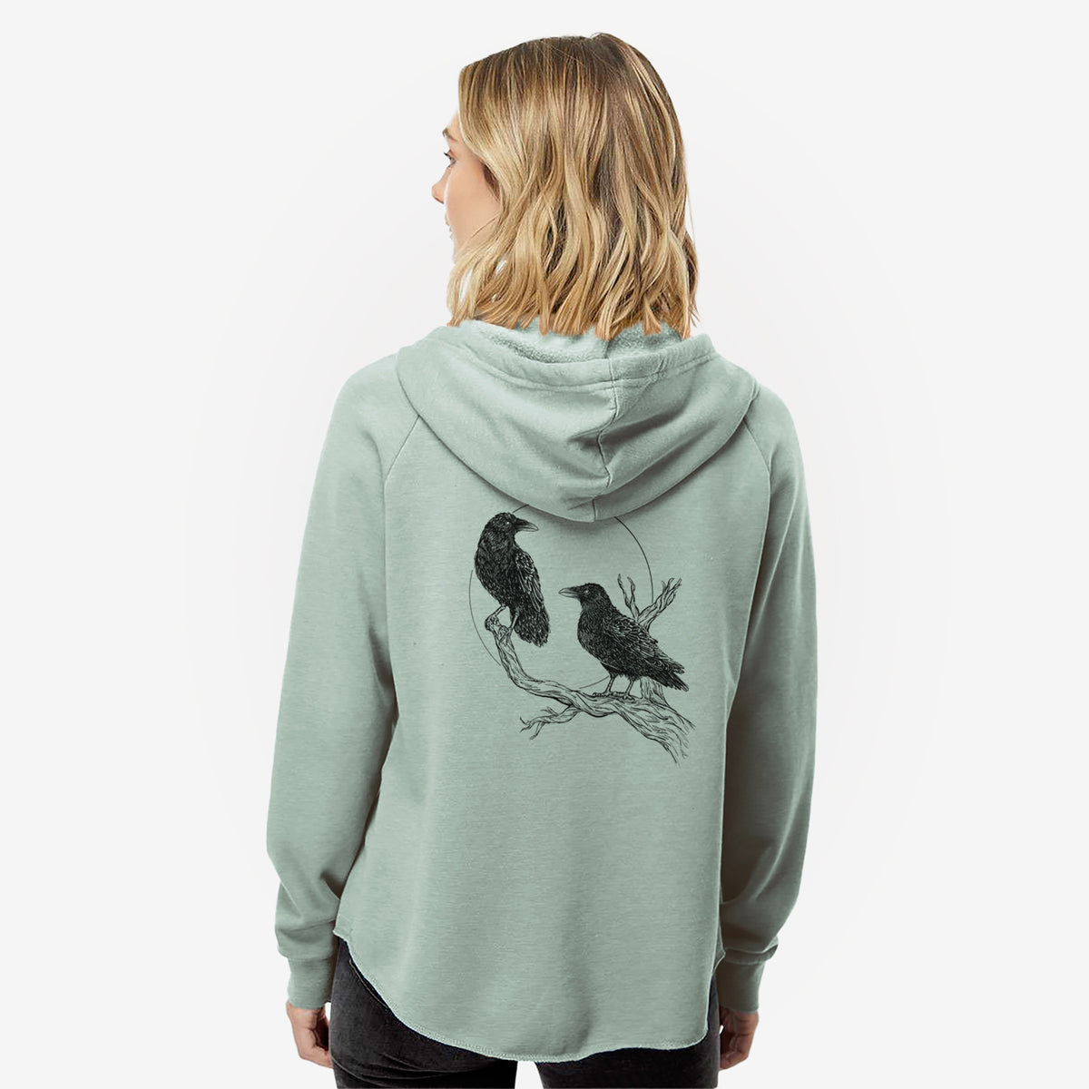 Woman wearing a sage green women's Cali Wave zip-up sweatshirt printed with a black ink illustration of two ravens perched on a branch in front of a full moon, seen from behind.