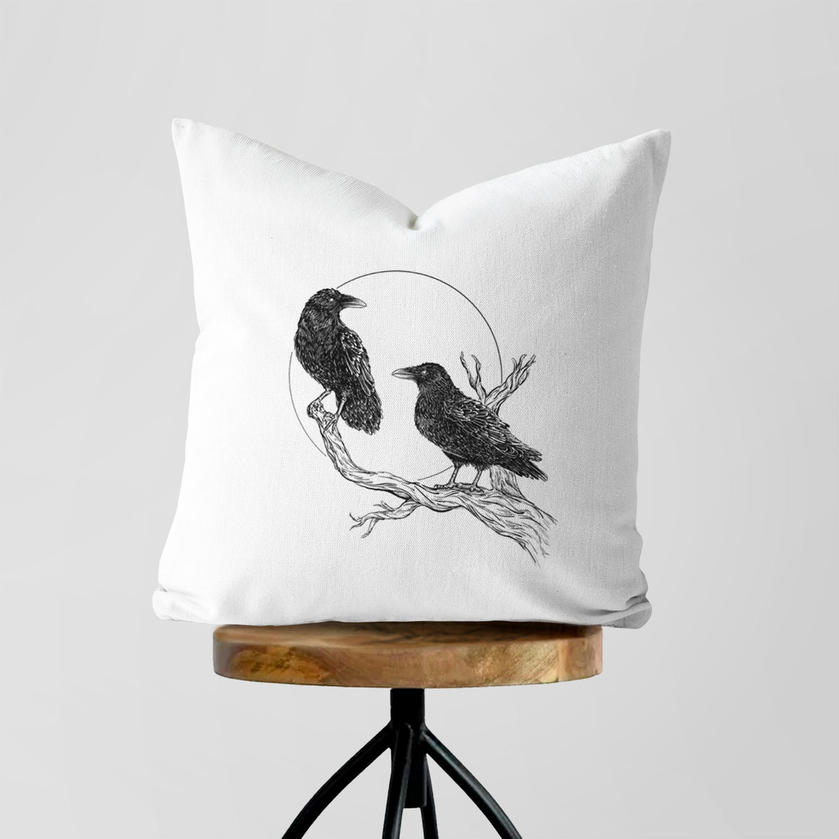 White pillow cover printed with a black ink illustration of two ravens perched on a branch in front of a full moon, on a wooden stool.