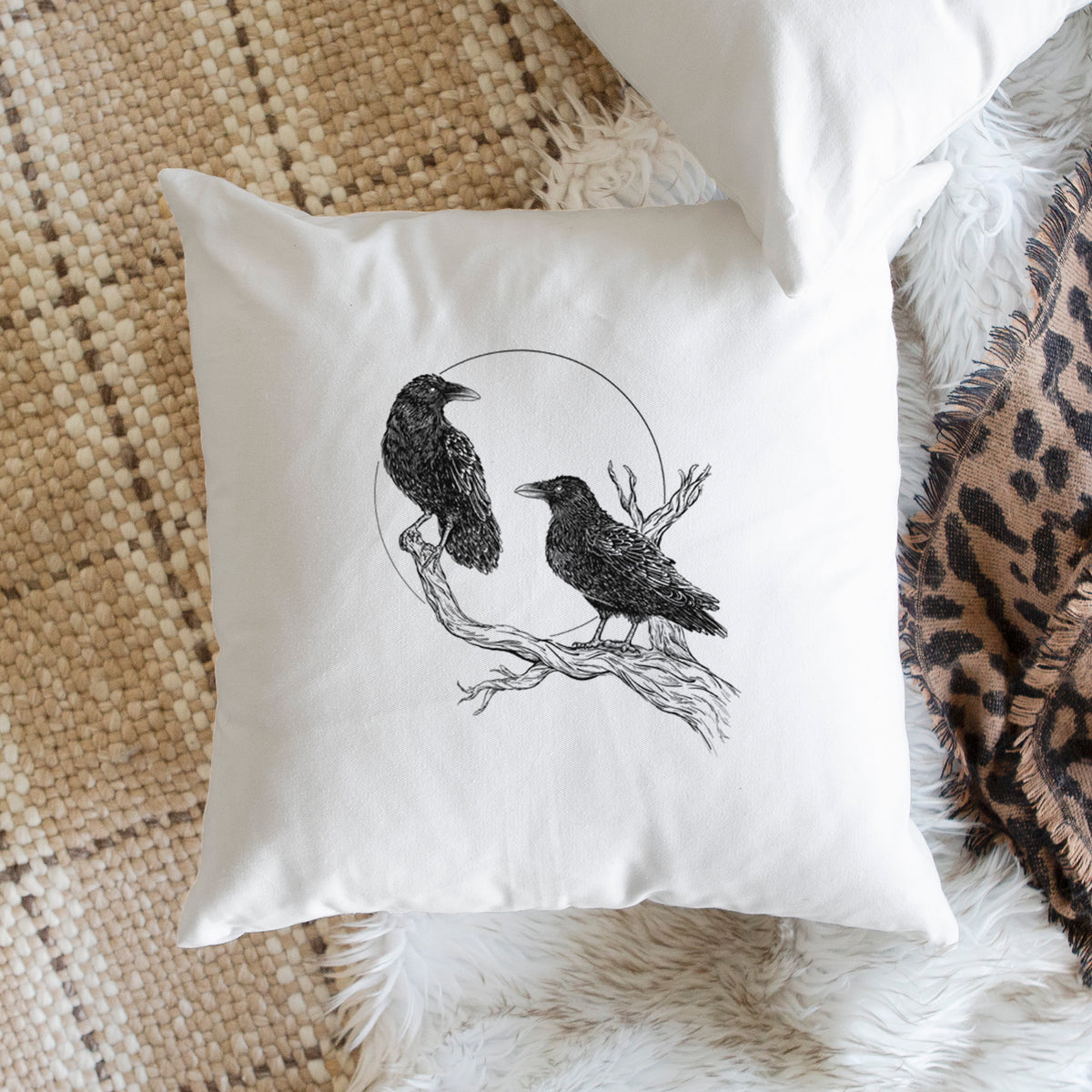 White pillow cover printed with a black ink illustration of two ravens perched on a branch in front of a full moon, on a woven chair.