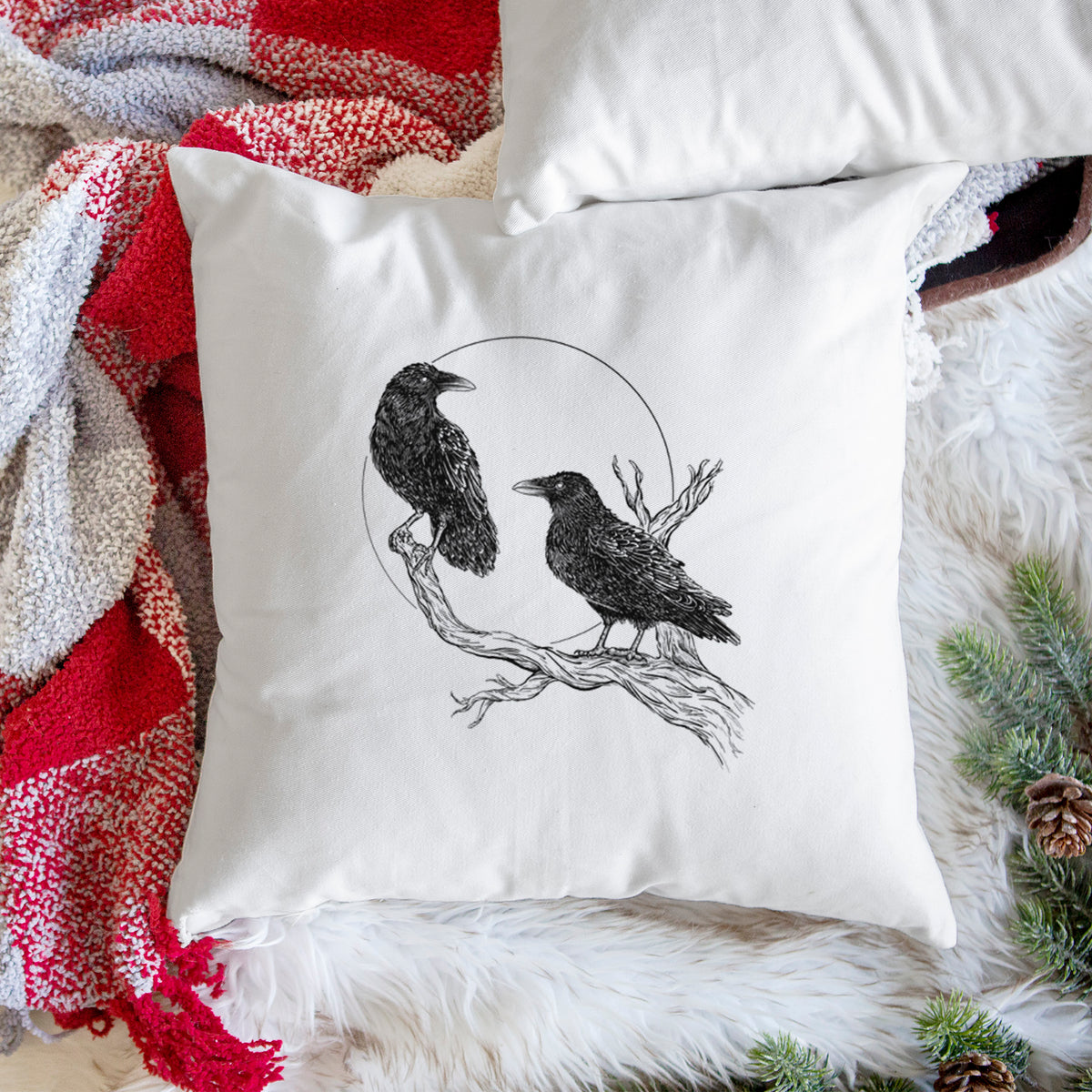 White pillow cover printed with a black ink illustration of two ravens perched on a branch in front of a full moon, on a woven chair.