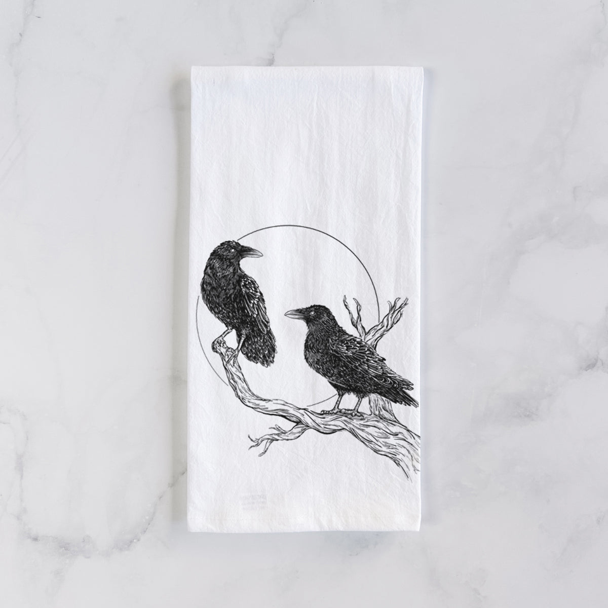 White cotton tea towel laid flat on marble, printed with a black ink illustration of two ravens perched on a branch in front of a full moon.
