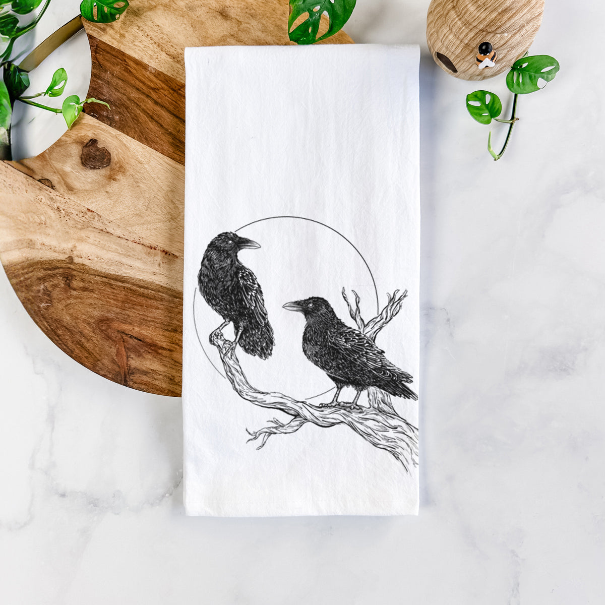White cotton tea towel printed with a black ink illustration of two ravens perched on a branch in front of a full moon, on a marble counter beside a round wooden cutting board and greenery.