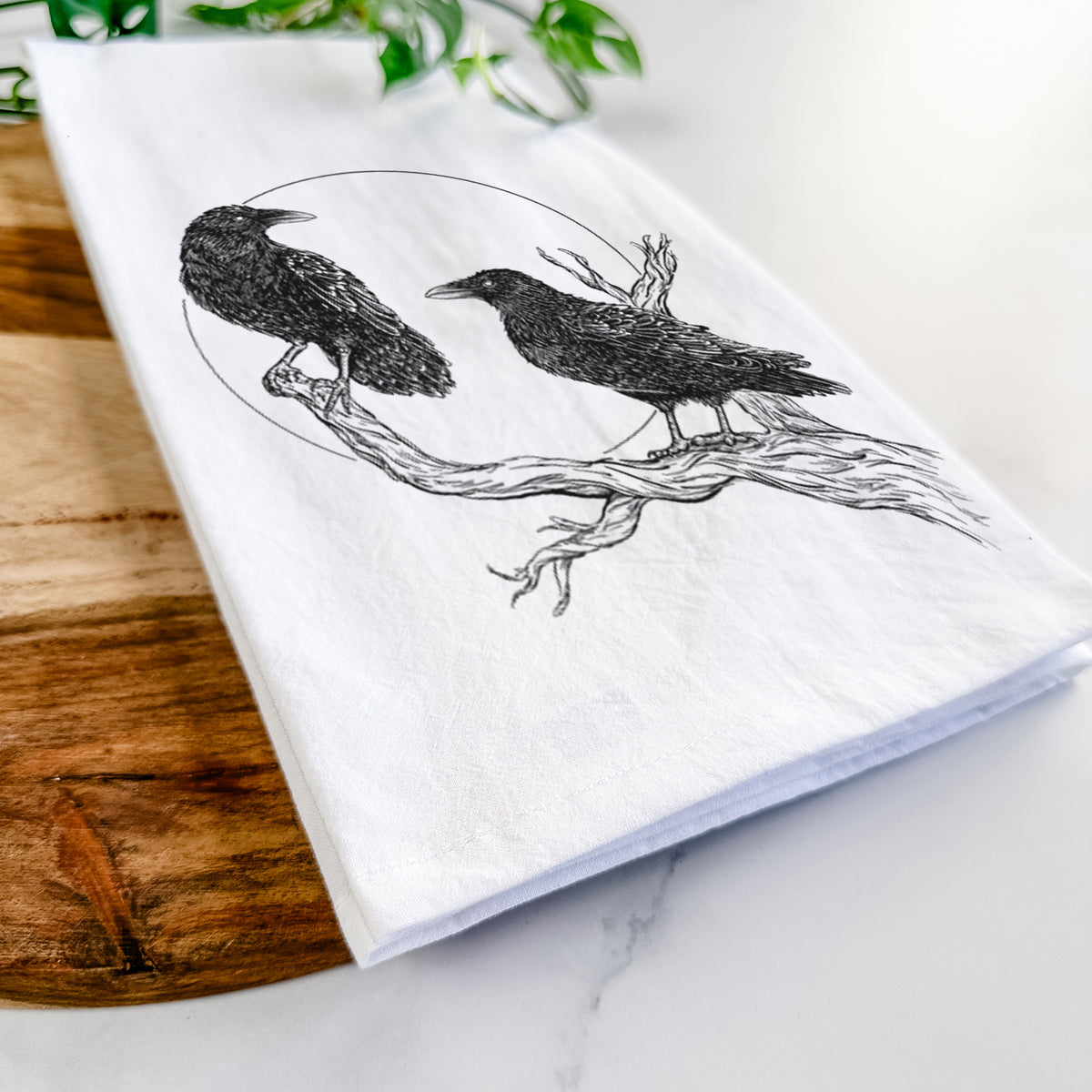 Close-up of a folded white tea towel on a wooden board, showing a black ink illustration of two ravens perched on a branch in front of a full moon.