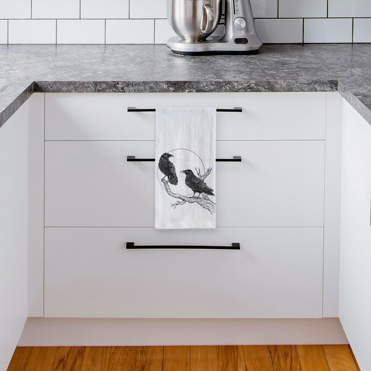 White tea towel printed with a black ink illustration of two ravens perched on a branch in front of a full moon, hanging from a drawer handle in a bright white kitchen.