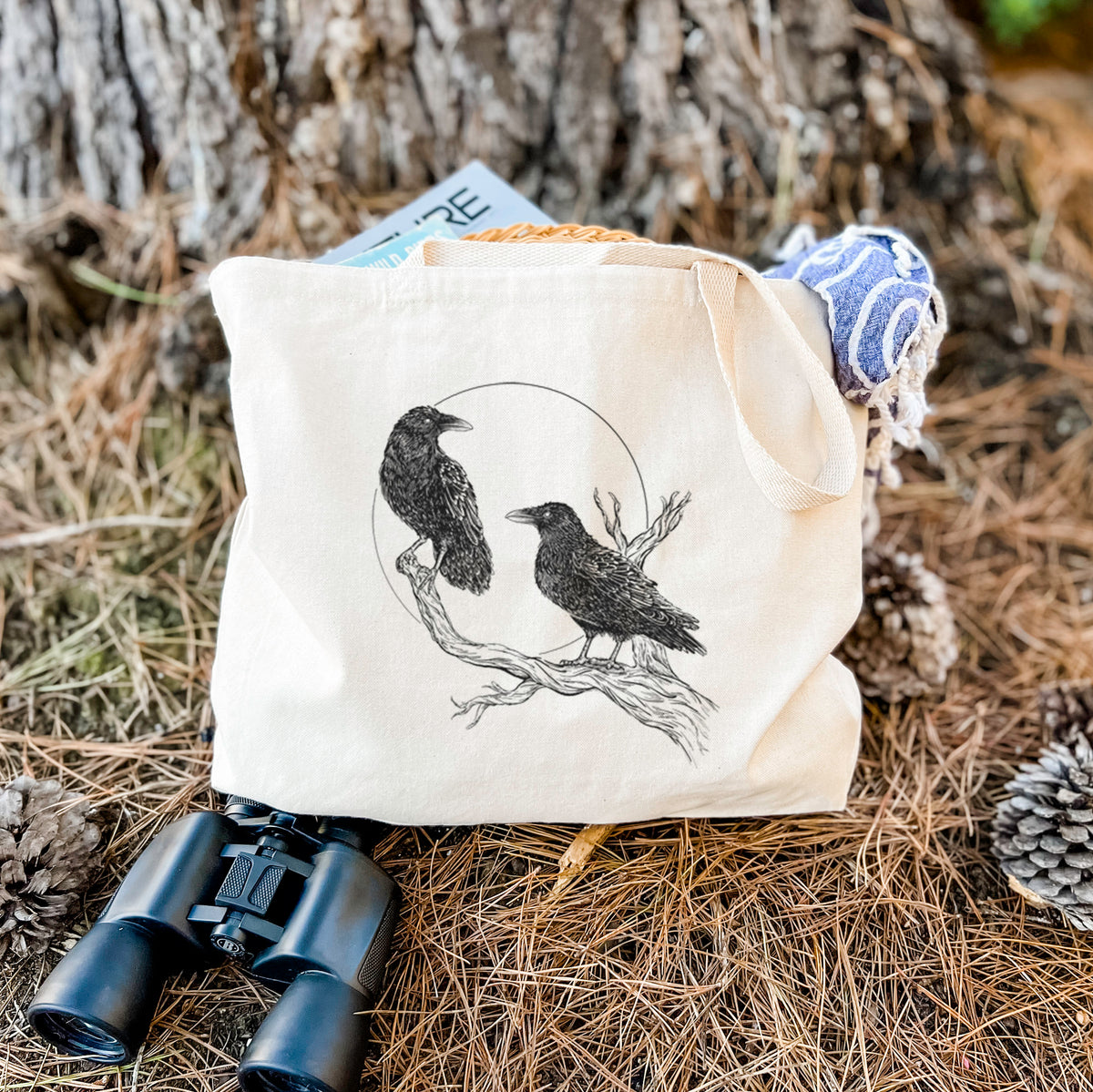 Canvas tote bag printed with a black ink illustration of two ravens perched on a branch in front of a full moon, resting on dry grass beside a pair of binoculars.