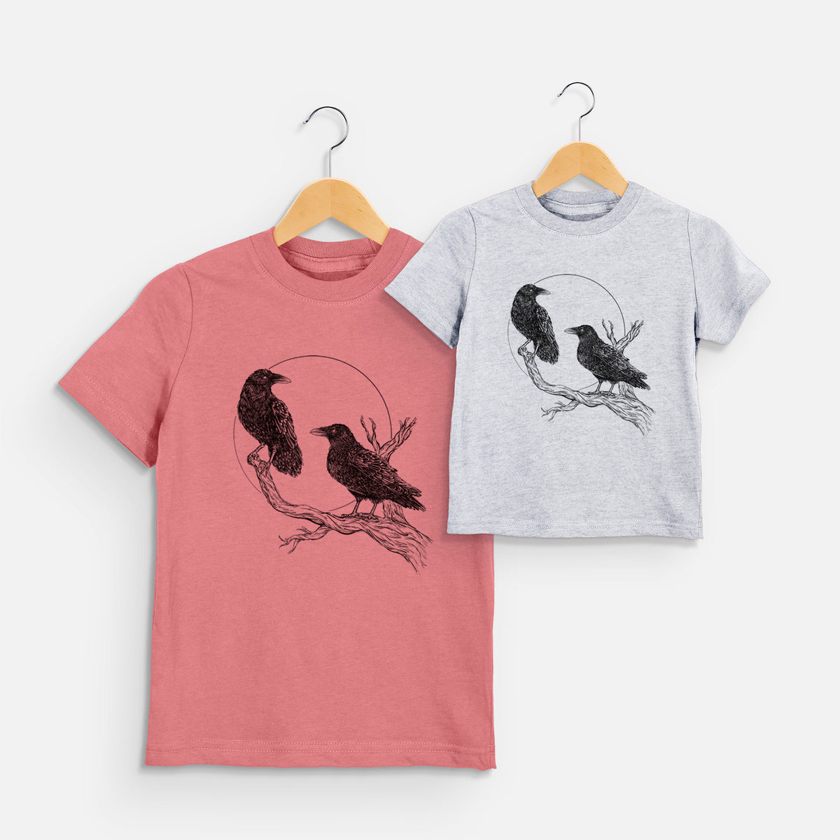 Two youth t-shirts in different colors on hangers, printed with a black ink illustration of two ravens perched on a branch in front of a full moon.