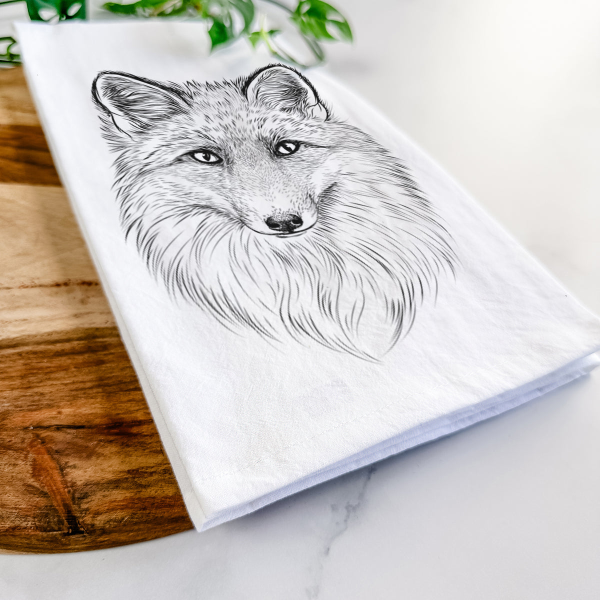 Close-up of a folded white tea towel on a wooden board, showing a detailed black ink sketch of a red fox's face looking straight ahead, its fur drawn in fine pencil-like strokes.
