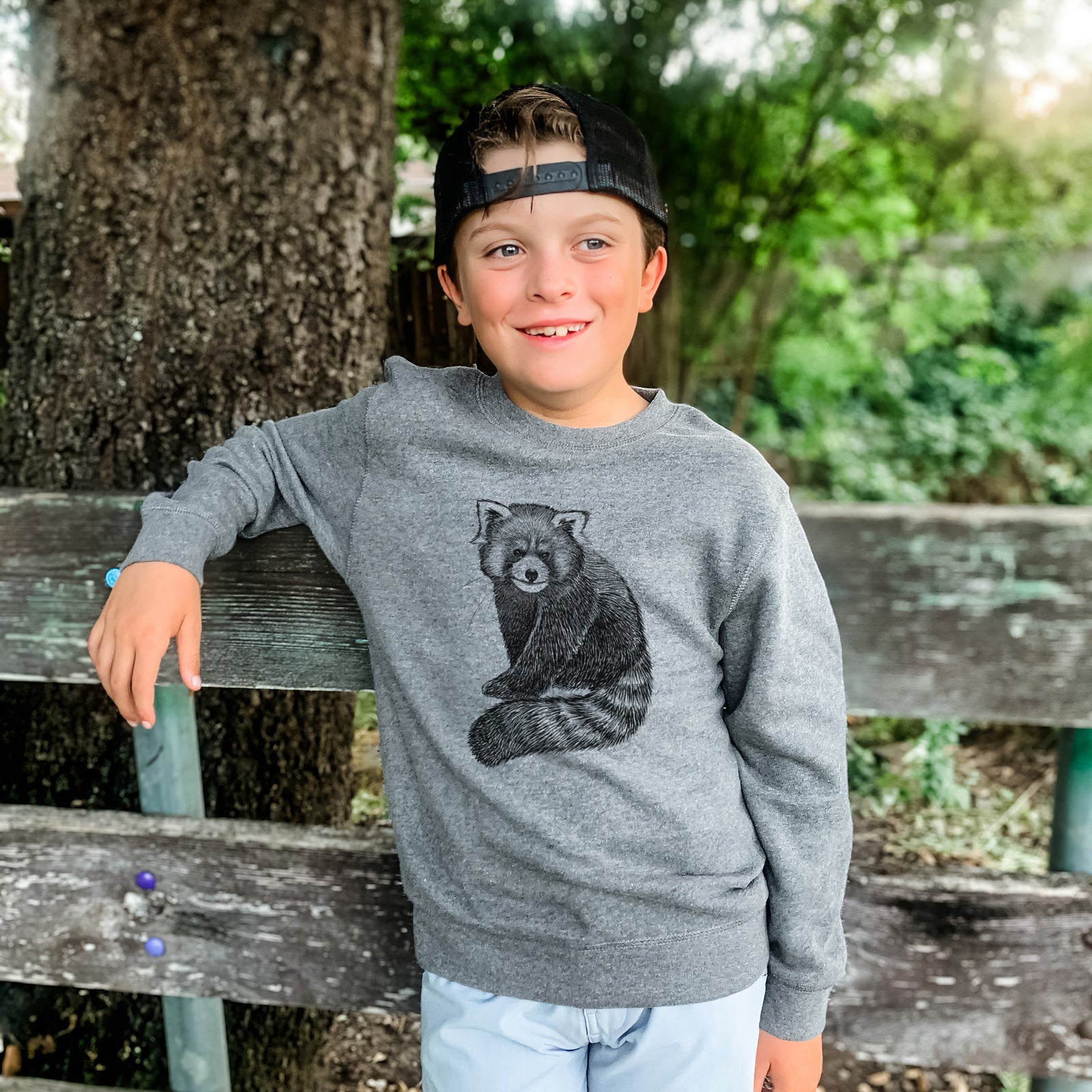 Boy wearing a youth crewneck sweatshirt printed with a detailed black ink illustration of a red panda sitting with its striped tail wrapped around, outdoors.