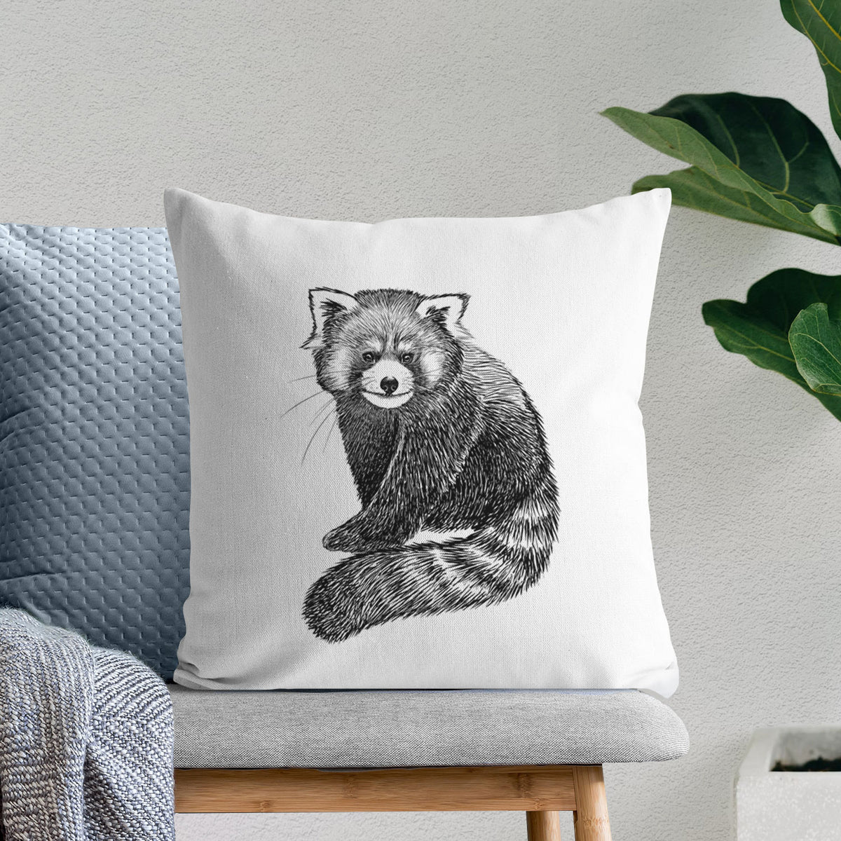 White pillow cover printed with a detailed black ink illustration of a red panda sitting with its striped tail wrapped around, on a bench beside plants.