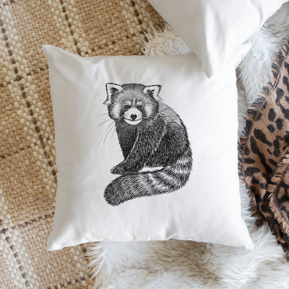 White pillow cover printed with a detailed black ink illustration of a red panda sitting with its striped tail wrapped around, on a woven chair.