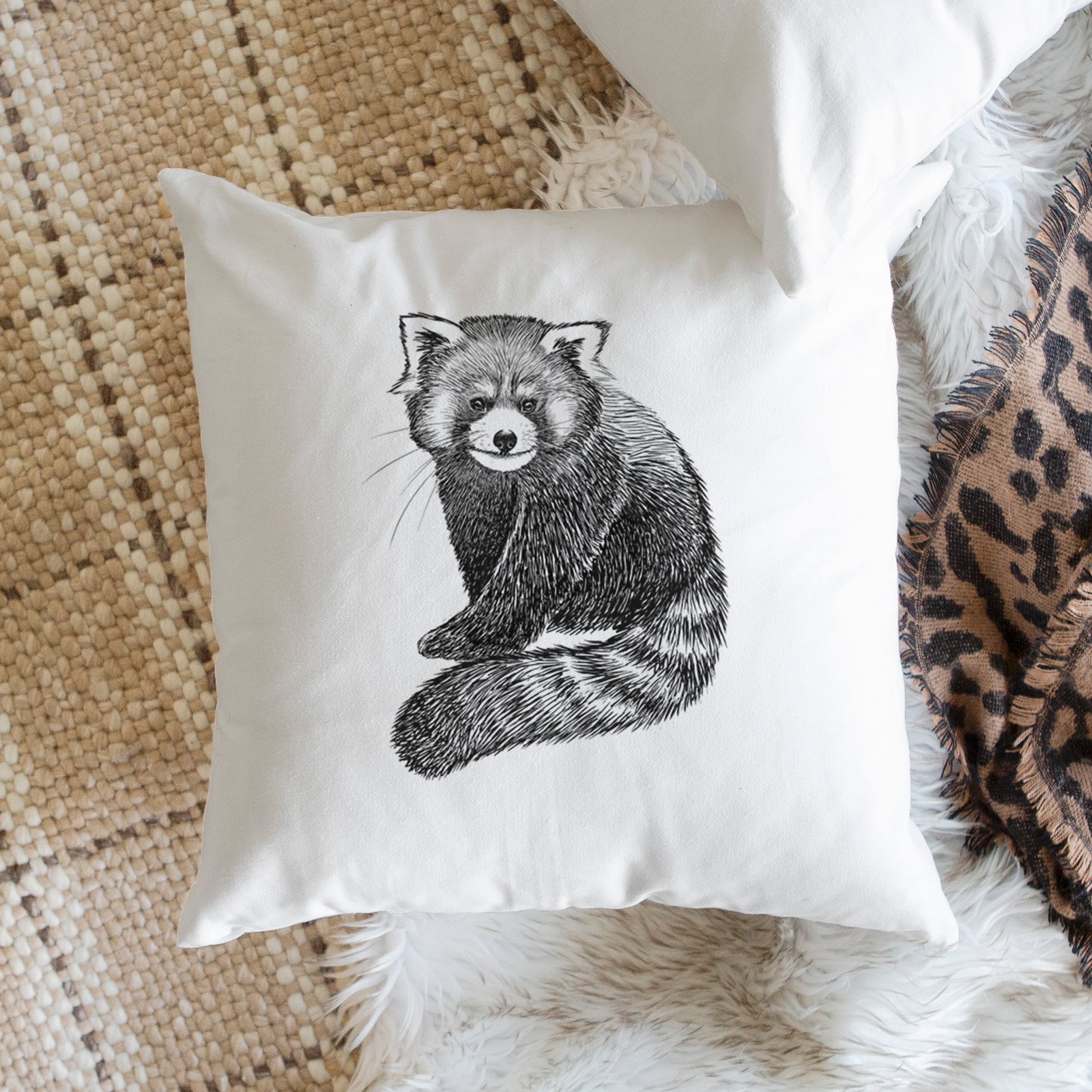 White pillow cover printed with a detailed black ink illustration of a red panda sitting with its striped tail wrapped around, on a bench beside plants.