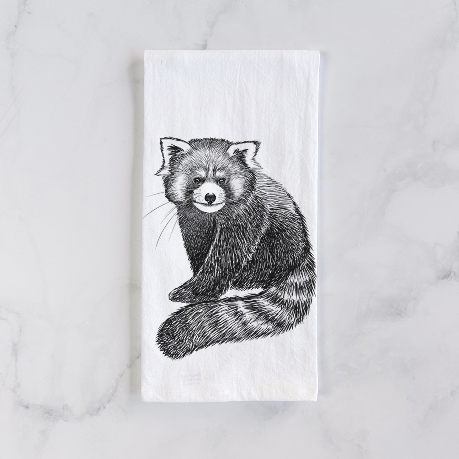 White cotton tea towel printed with a detailed black ink illustration of a red panda sitting with its striped tail wrapped around, on a marble counter beside a round wooden cutting board and greenery.