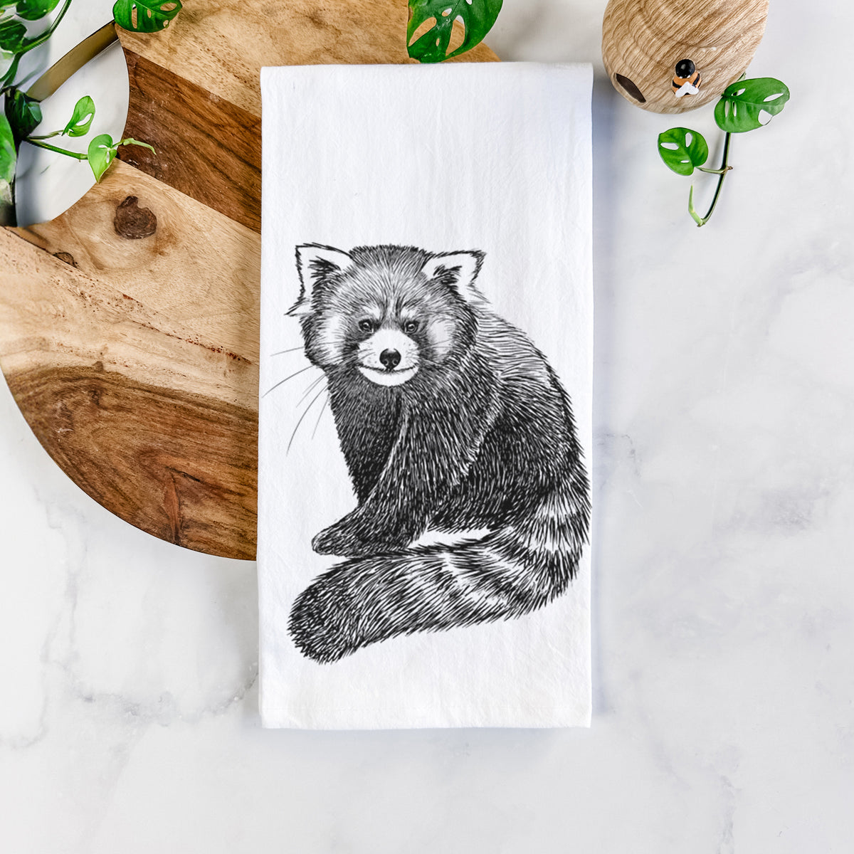 White cotton tea towel printed with a detailed black ink illustration of a red panda sitting with its striped tail wrapped around, on a marble counter beside a round wooden cutting board and greenery.