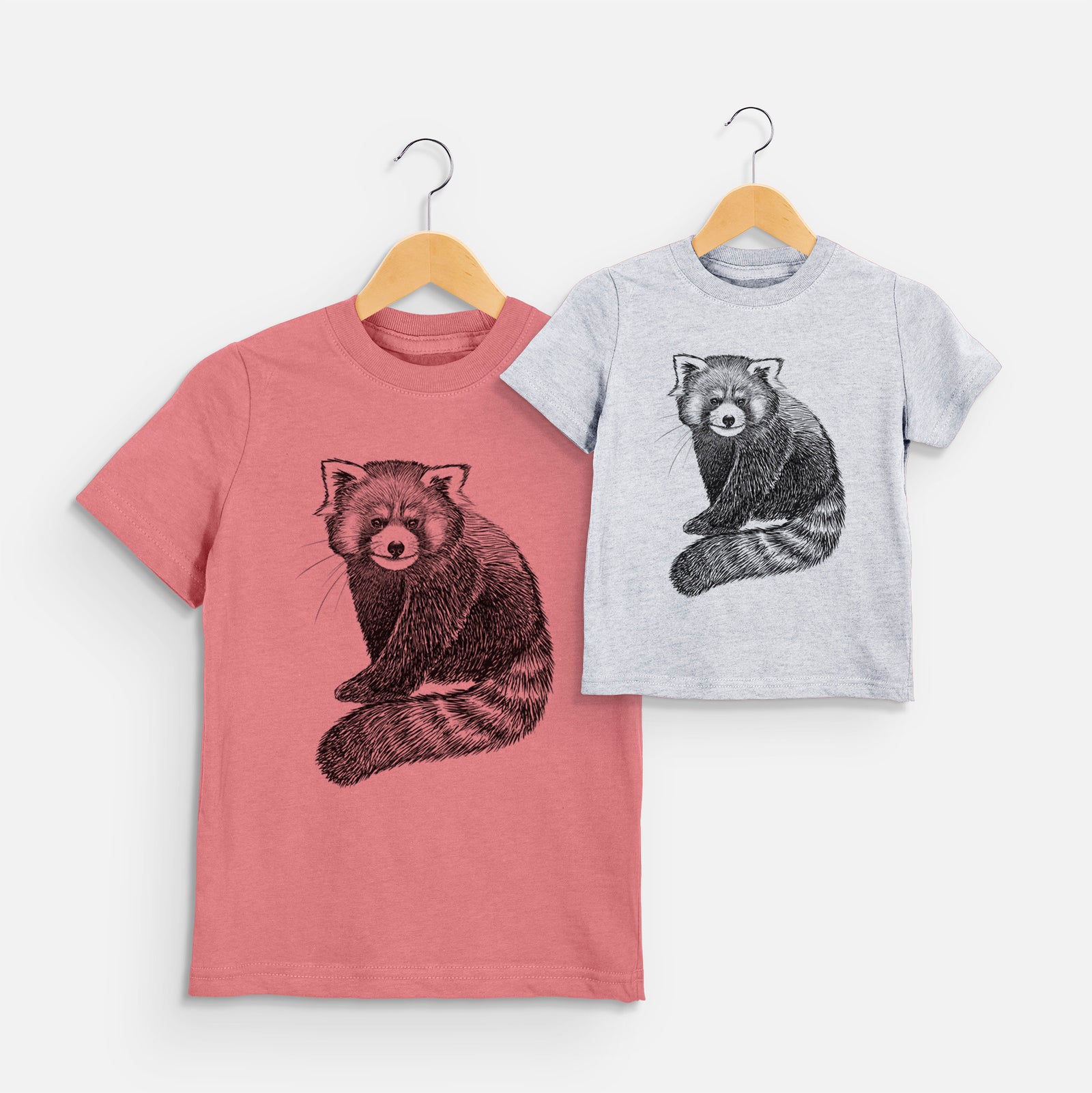 Two youth t-shirts in different colors on hangers, printed with a detailed black ink illustration of a red panda sitting with its striped tail wrapped around.