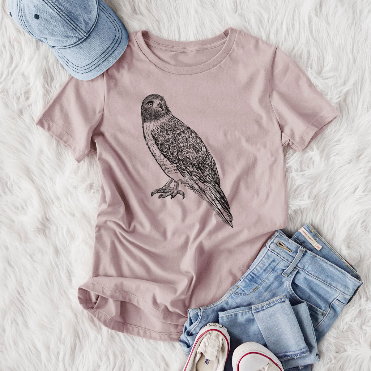 Women's relaxed fit cotton t-shirt printed with a detailed black ink illustration of a red-tailed hawk perched, styled flat on a white faux fur rug with jeans.