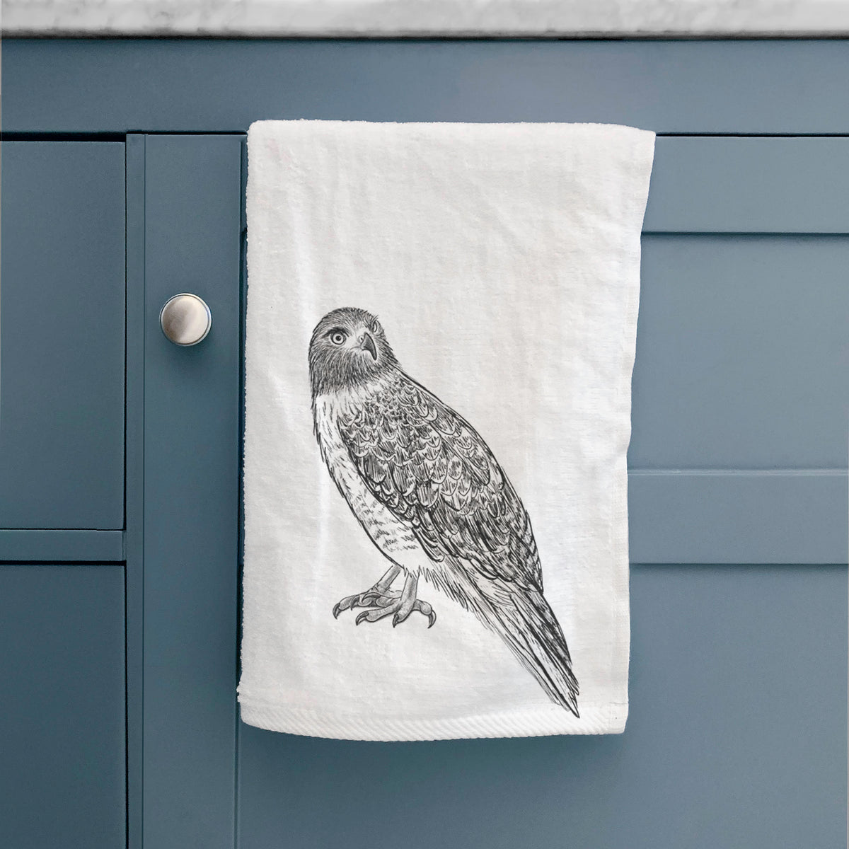 White hand towel printed with a detailed black ink illustration of a red-tailed hawk perched, hanging on a cabinet door.
