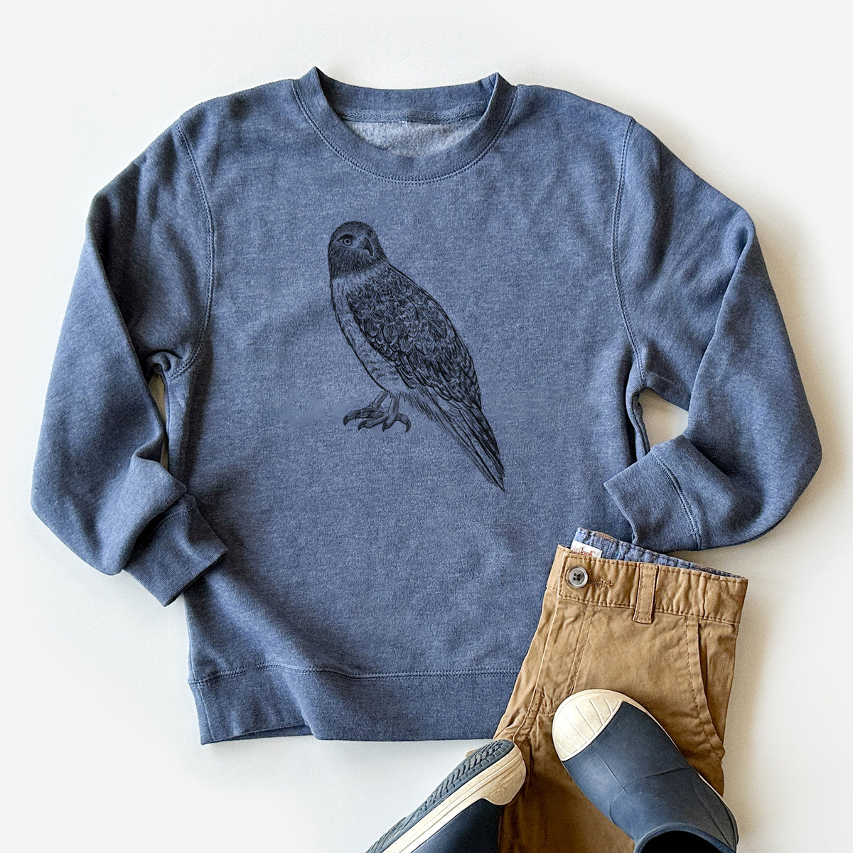 Youth crewneck sweatshirt printed with a detailed black ink illustration of a red-tailed hawk perched, styled flat with khaki pants and shoes.