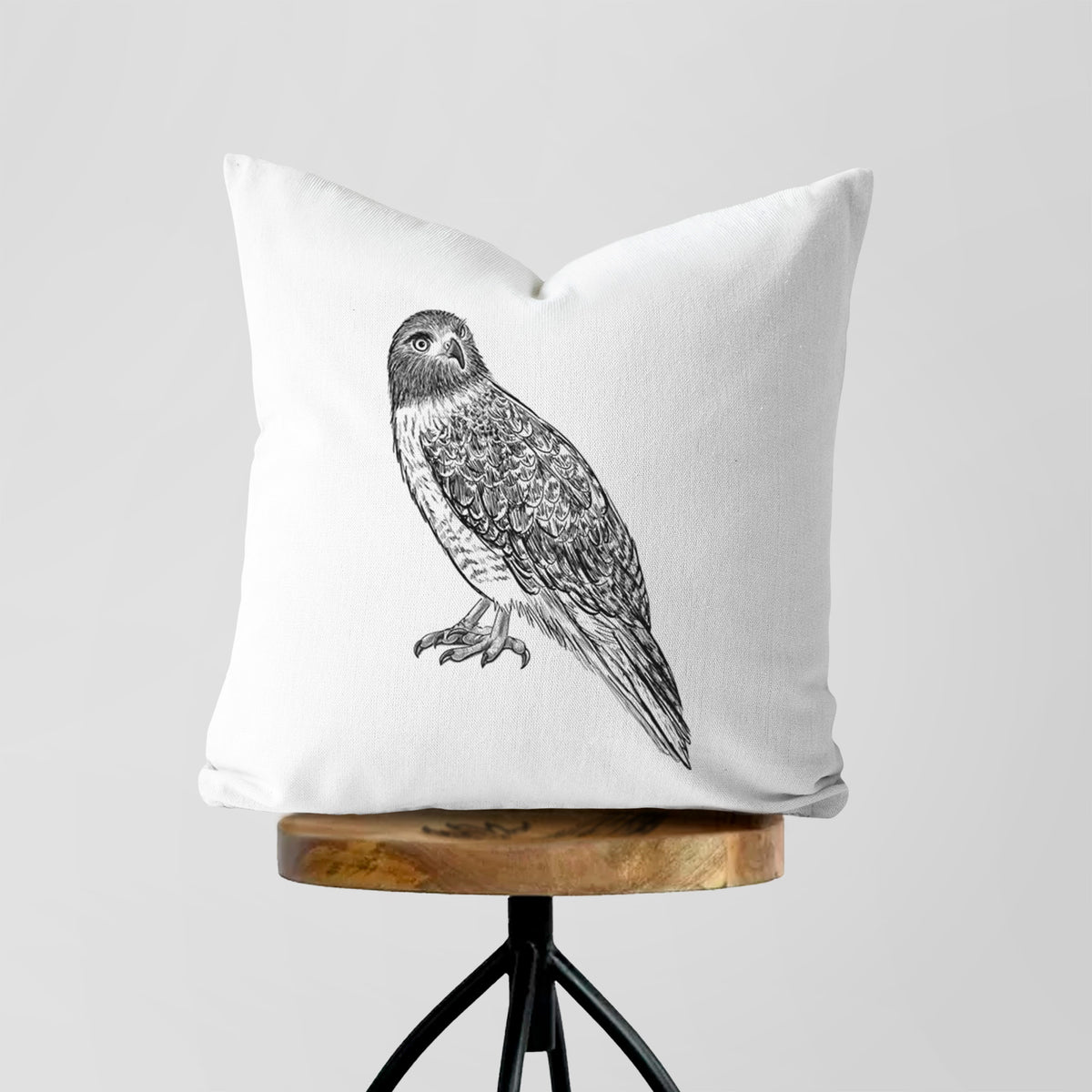 White pillow cover printed with a detailed black ink illustration of a red-tailed hawk perched, on a wooden stool.
