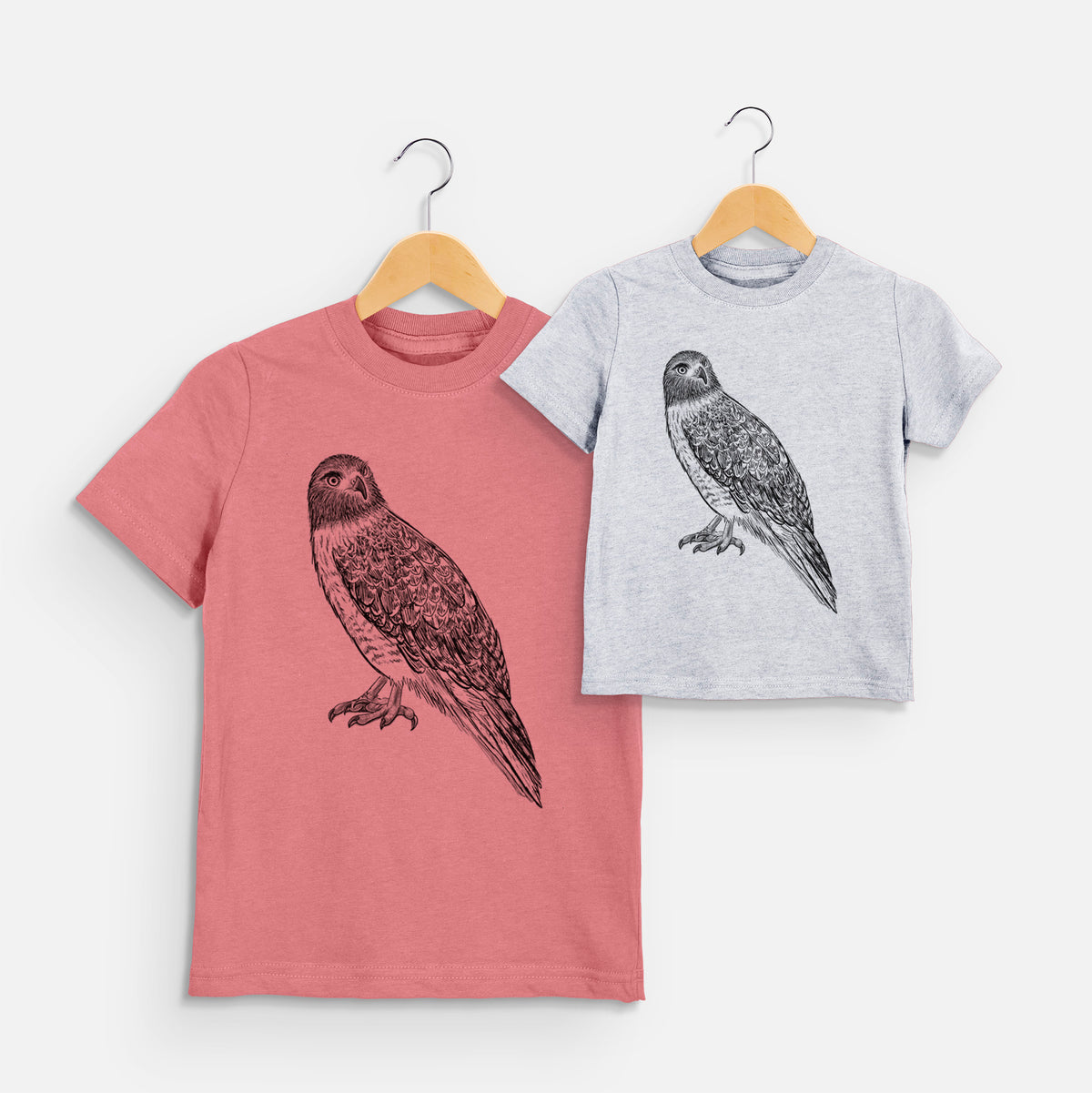 Two youth t-shirts in different colors on hangers, printed with a detailed black ink illustration of a red-tailed hawk perched.