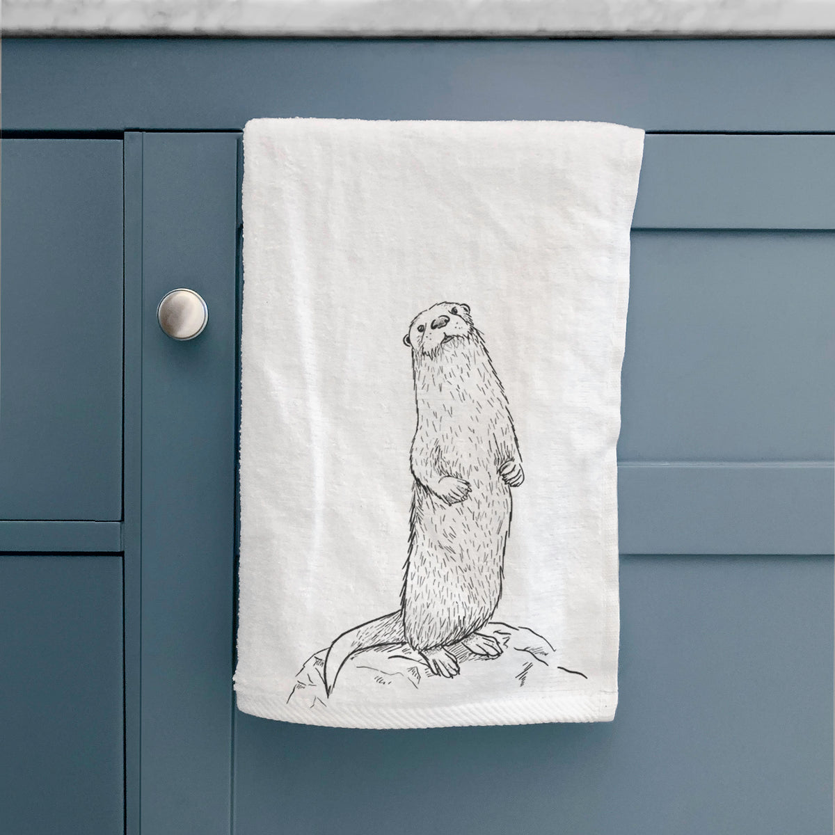 White hand towel printed with a black ink illustration of a river otter standing upright on a rock, hanging on a cabinet door.
