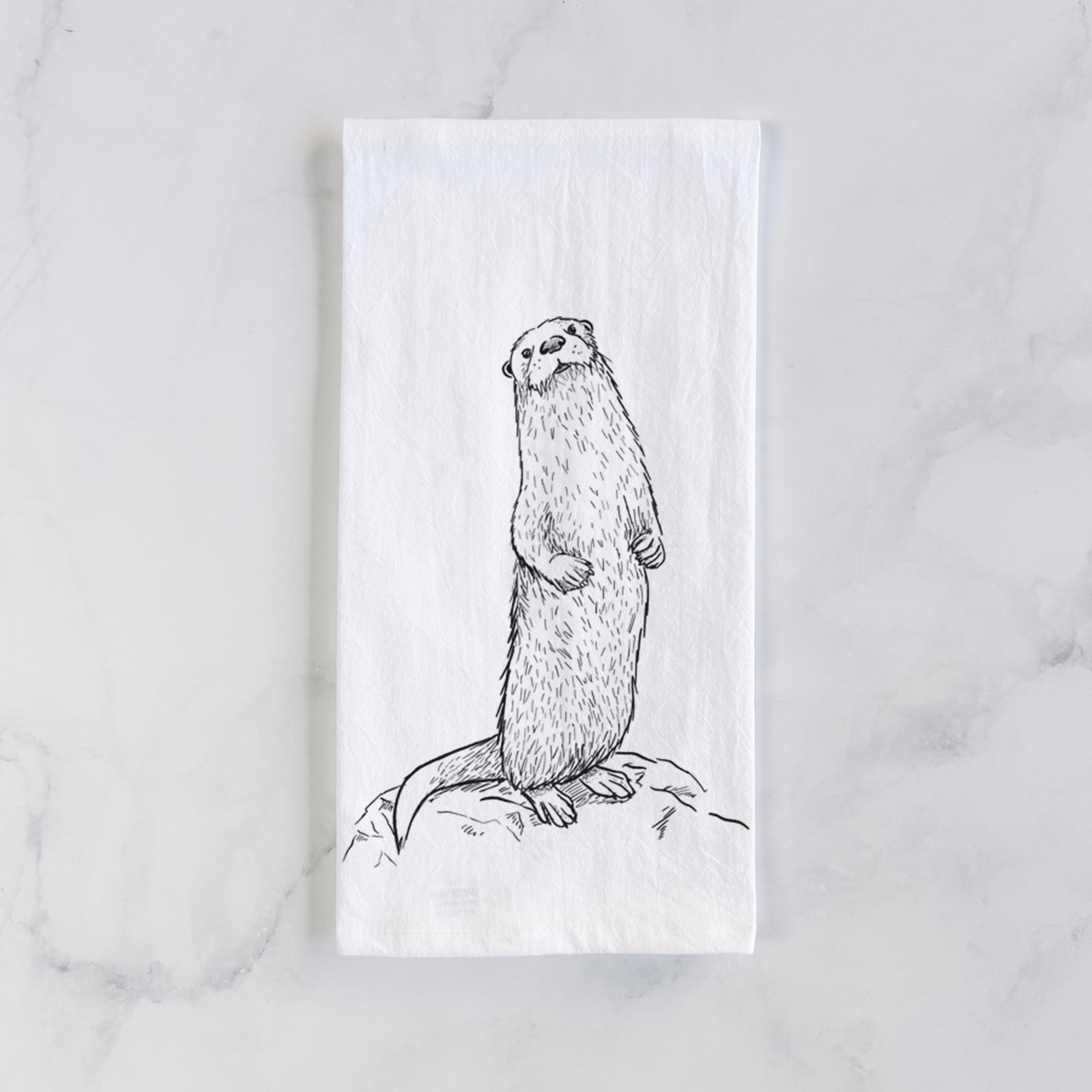 White cotton tea towel printed with a black ink illustration of a river otter standing upright on a rock, on a marble counter beside a round wooden cutting board and greenery.