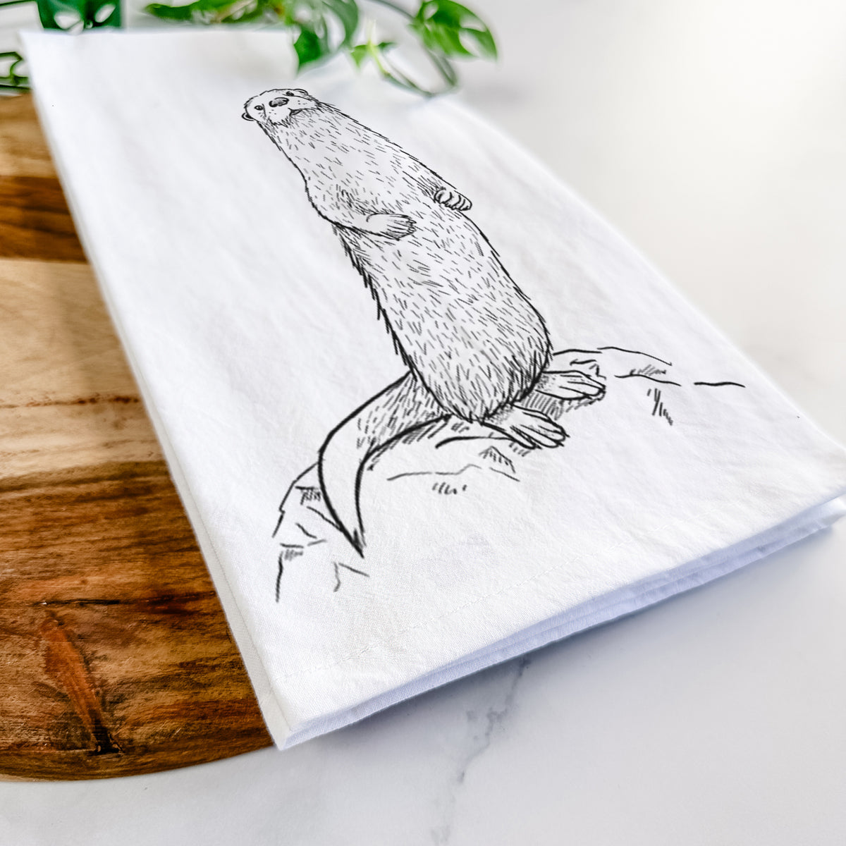 Close-up of a folded white tea towel on a wooden board, showing a black ink illustration of a river otter standing upright on a rock.