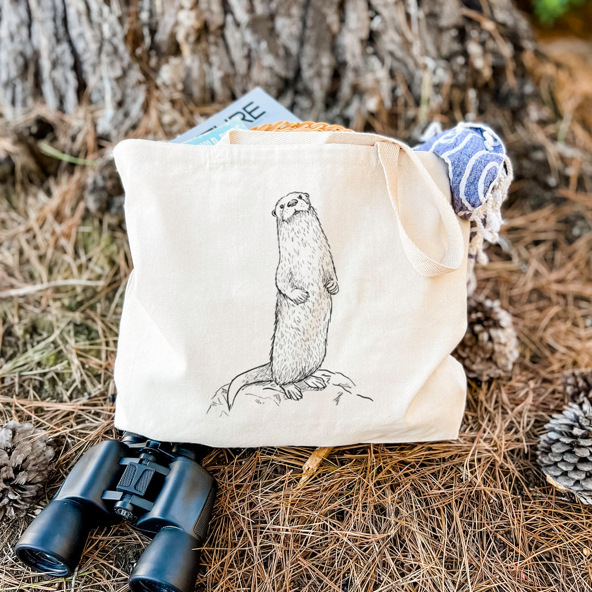 Canvas tote bag printed with a black ink illustration of a river otter standing upright on a rock, resting on dry grass beside a pair of binoculars.