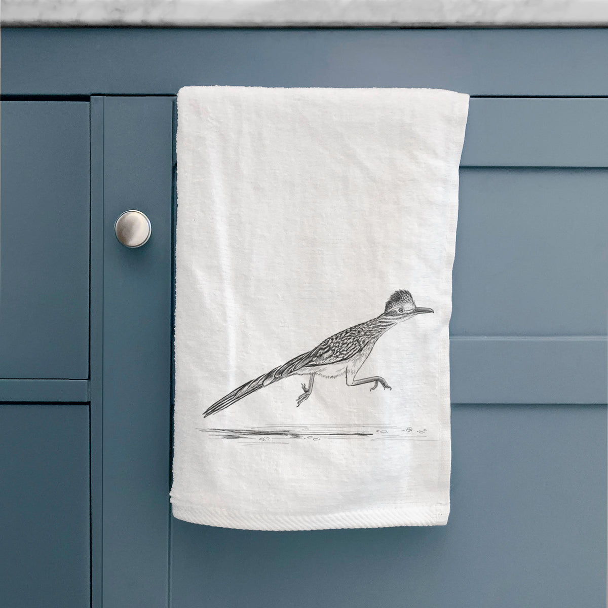 White hand towel printed with a black ink illustration of a roadrunner running, hanging on a cabinet door.