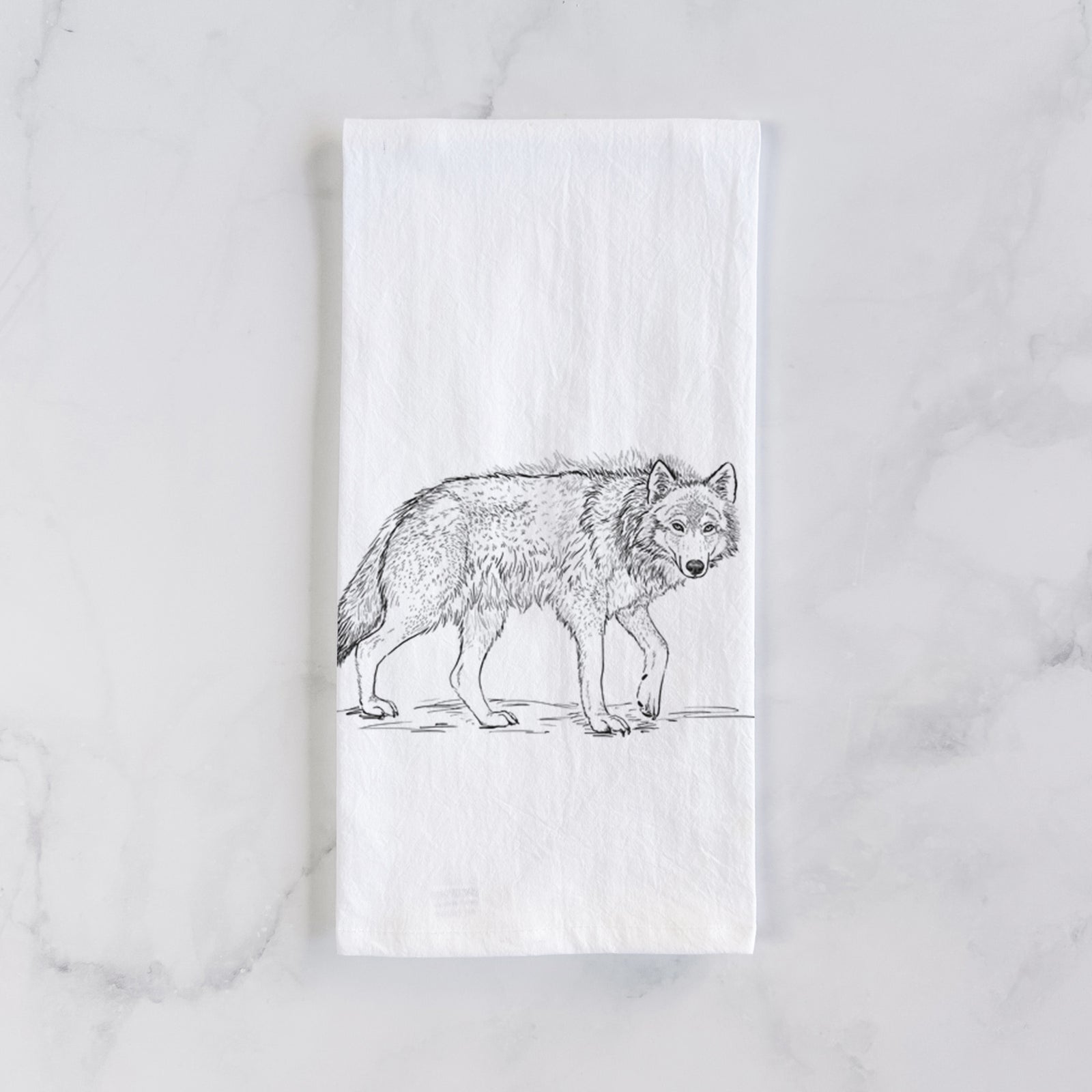 White cotton tea towel printed with a detailed black ink illustration of a grey wolf walking, on a marble counter beside a round wooden cutting board and greenery.