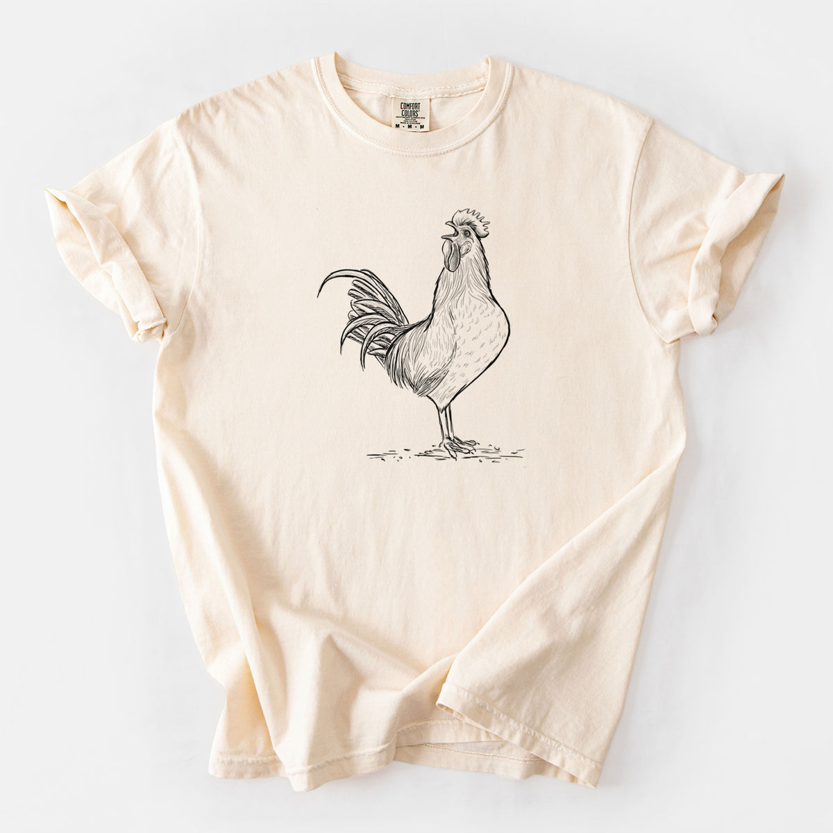Ivory Comfort Colors t-shirt laid flat, printed with a detailed black ink illustration of a rooster crowing.